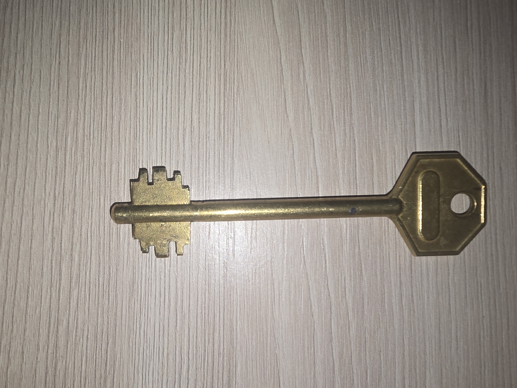A brass key lies vertically on a light-colored wooden surface. The key has a hexagonal head with a circular hole and an elongated oval indentation. The shaft of the key is thin and cylindrical, leading to a bit with two notches on each side, resembling a comb. The wooden surface has a distinct grain pattern. There are no people or actions depicted in the image. The location in Ulan-Ude, Russia, is not determinable from the visual information provided.