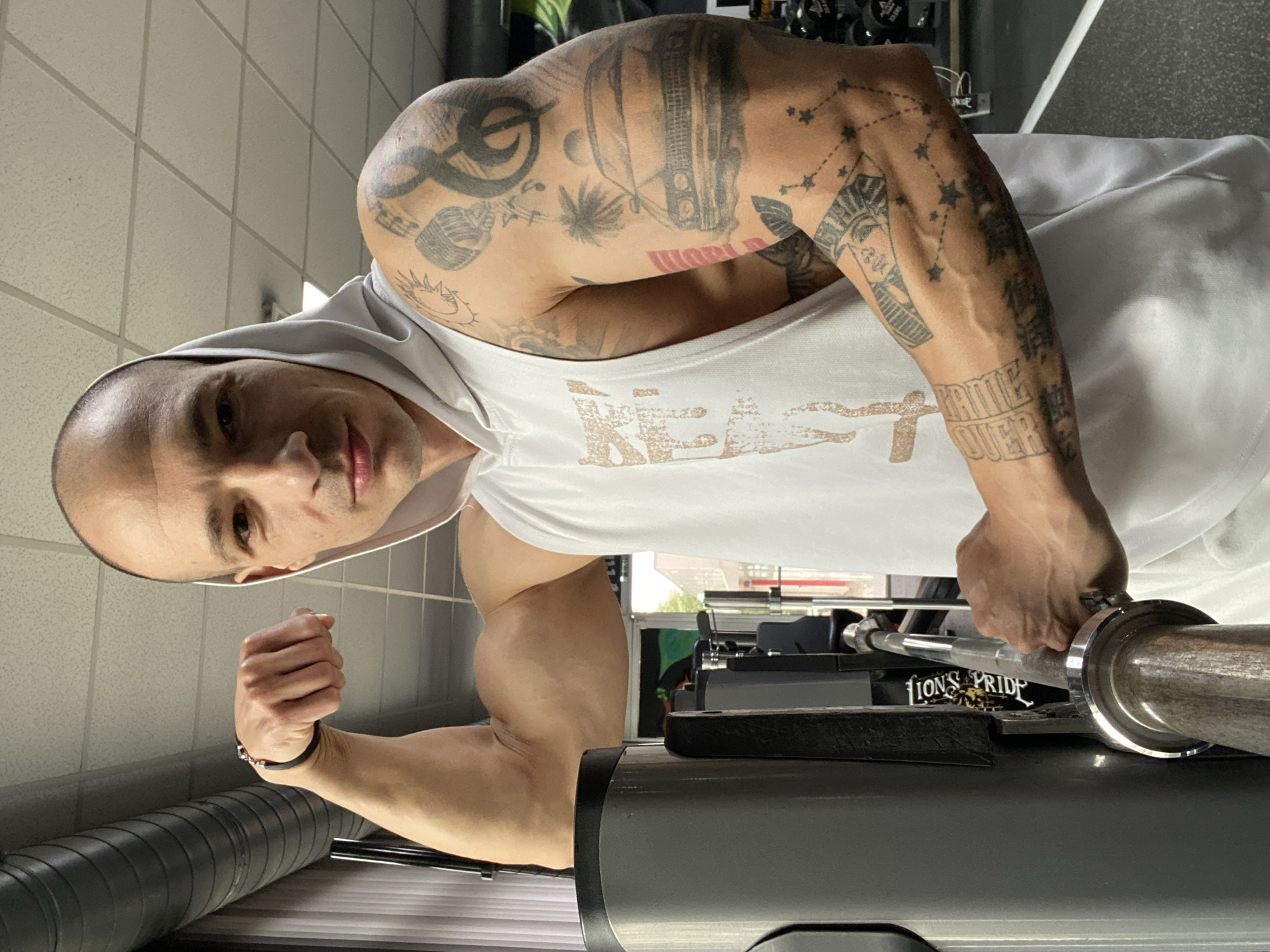 A bald man with extensive tattoos on his arms is in what appears to be a gym. He is wearing a white hooded tank top. His left arm is flexed, showing his bicep. His right hand is holding a barbell on a weight machine. The machine has the words "LIONS PRIDE" on it. The environment appears to be a well-lit gym with weights and workout equipment in the background. There is no identifiable information to suggest the location is Pleven, Bulgaria.