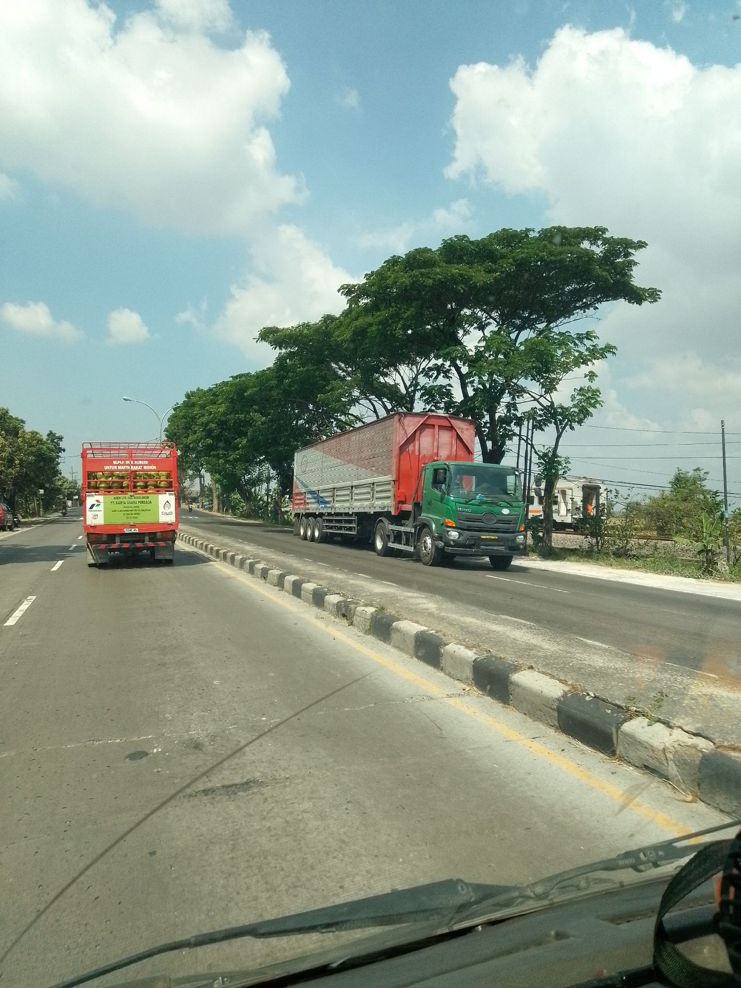 In Gembong, Indonesia, on a paved road under a partly cloudy sky, two trucks are traveling side by side. The truck on the left is red and carries yellow gas cylinders. The truck on the right is green and has a red trailer loaded with metal sheeting. The road has a yellow line marking the lane and a concrete curb with black and white bollards. Trees line the side of the road, and in the background, a train can be seen on tracks. The image is taken from a vehicle, as evidenced by the windshield wipers and the dashboard in the foreground.