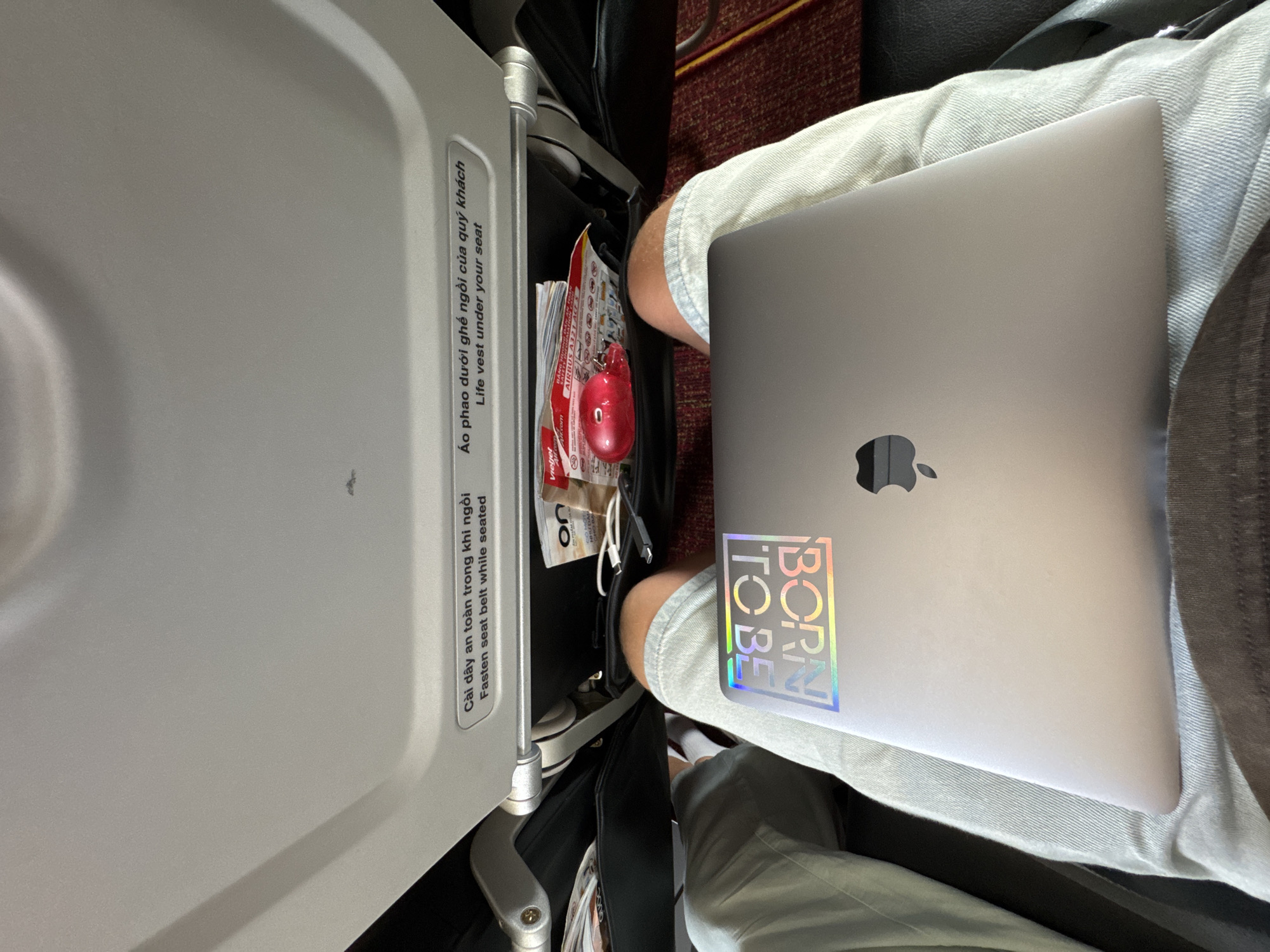 The image captures an overhead view of a person's lap and the airplane cabin in front of them. A silver MacBook is open and resting on the person's thighs. A holographic sticker reading "BORN TO BE" in a stylized font is adhered to the lid of the MacBook. The person is wearing light-wash denim shorts and a dark fabric garment is visible at the bottom of the frame.
In the background, the seatback pocket of the airplane seat in front is visible, containing various items including a red heart-shaped object, what appears to be a life vest, and some paper materials, possibly airline magazines or safety instructions. Text in both Vietnamese and English is present on the inside of the seatback, indicating instructions such as "Fasten seat belt while seated" and "Life vest under your seat." The overhead compartment of the airplane is also visible at the top of the image. The lighting suggests an indoor setting on an airplane. The location is identified as Thuận An, Vietnam, likely within the Tân Sơn Nhất International Airport, which serves the city.
