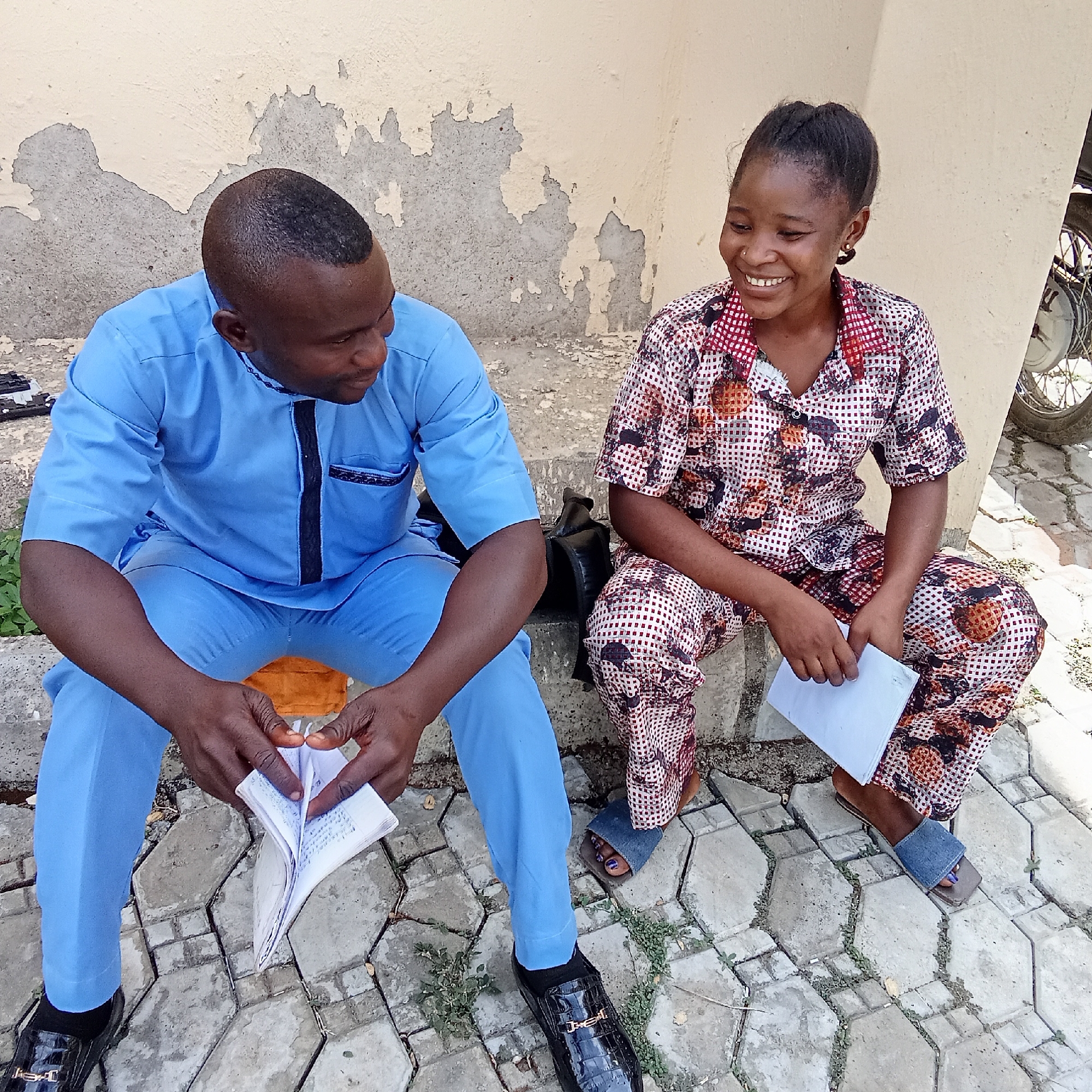 Two young adults, a man and a woman, are seated outdoors on a concrete ledge in Akwanga, Nigeria, engaged in a cheerful interaction. The man, positioned on the left, has dark skin and short hair, wearing a light blue traditional tunic with black accents and matching trousers. He sports black shiny loafers with a gold buckle that has partially legible text, possibly "JES". He holds an open notebook or small book, appearing to examine its contents while smiling subtly.
To his right, the woman has dark skin and her dark hair is pulled back. She is dressed in a vibrant two-piece outfit featuring a checkerboard and abstract pattern in white, red, and brown. She has a nose piercing and small earrings, and her toes are painted with purple nail polish, visible as she wears blue denim slide sandals. She holds white papers in her left hand and is looking towards the man, laughing heartily with a wide, joyful smile.
They are seated on a low concrete structure, with a textured grey interlocking block pavement beneath them, interspersed with small weeds. Behind them is a light-colored wall exhibiting significant wear and peeling paint. A black bag is visible near the man's left leg, and a portion of a motorcycle wheel can be seen in the upper right background. The scene is brightly lit, indicating daytime. Both individuals appear relaxed and happy, suggesting a friendly conversation or shared activity.