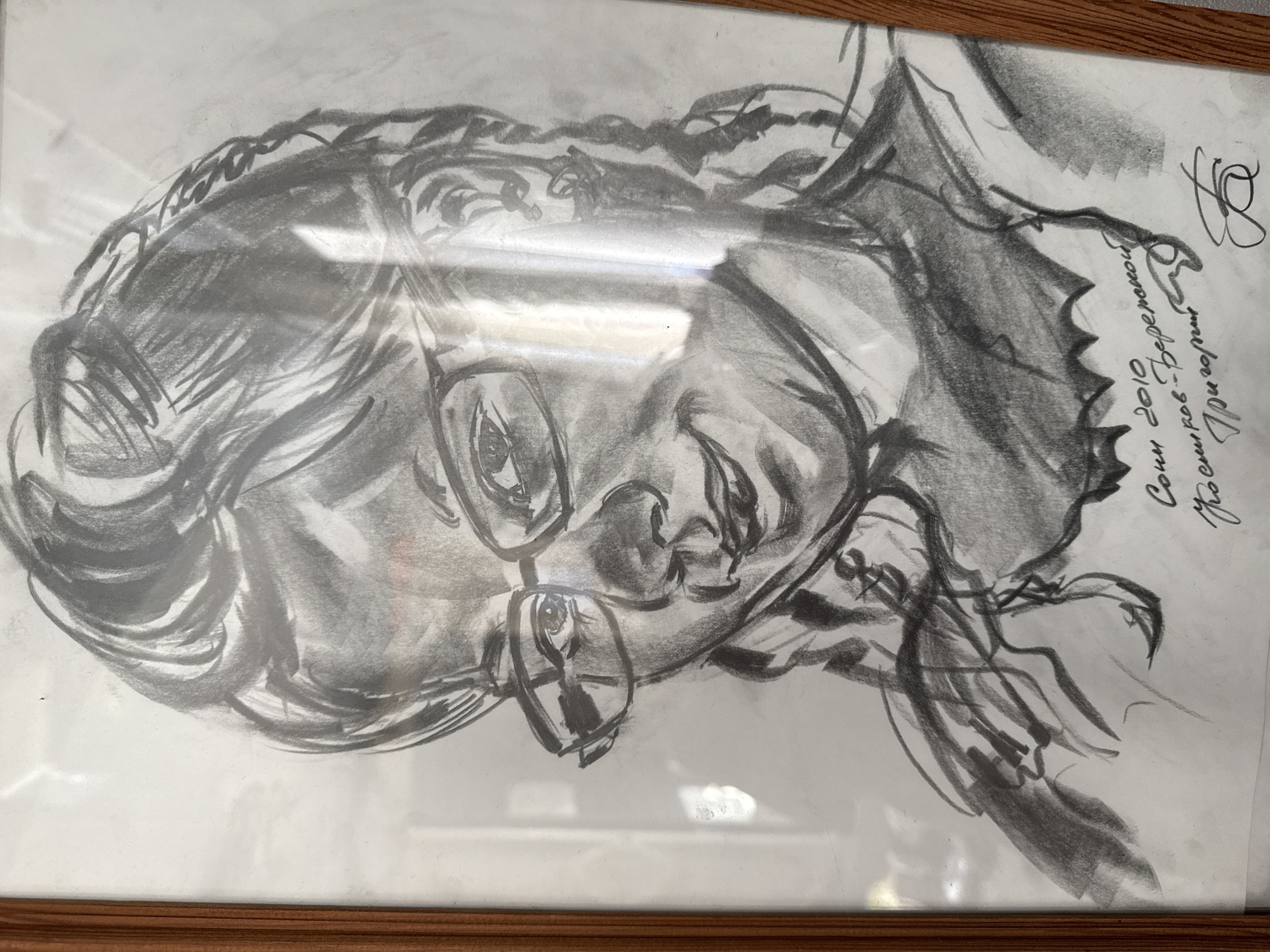 A framed charcoal drawing depicting a woman with glasses. In the upper right corner of the drawing, there is a signature with the words "Сочи 2010" and "Чайковский-Пермский". The frame is made of wood.
