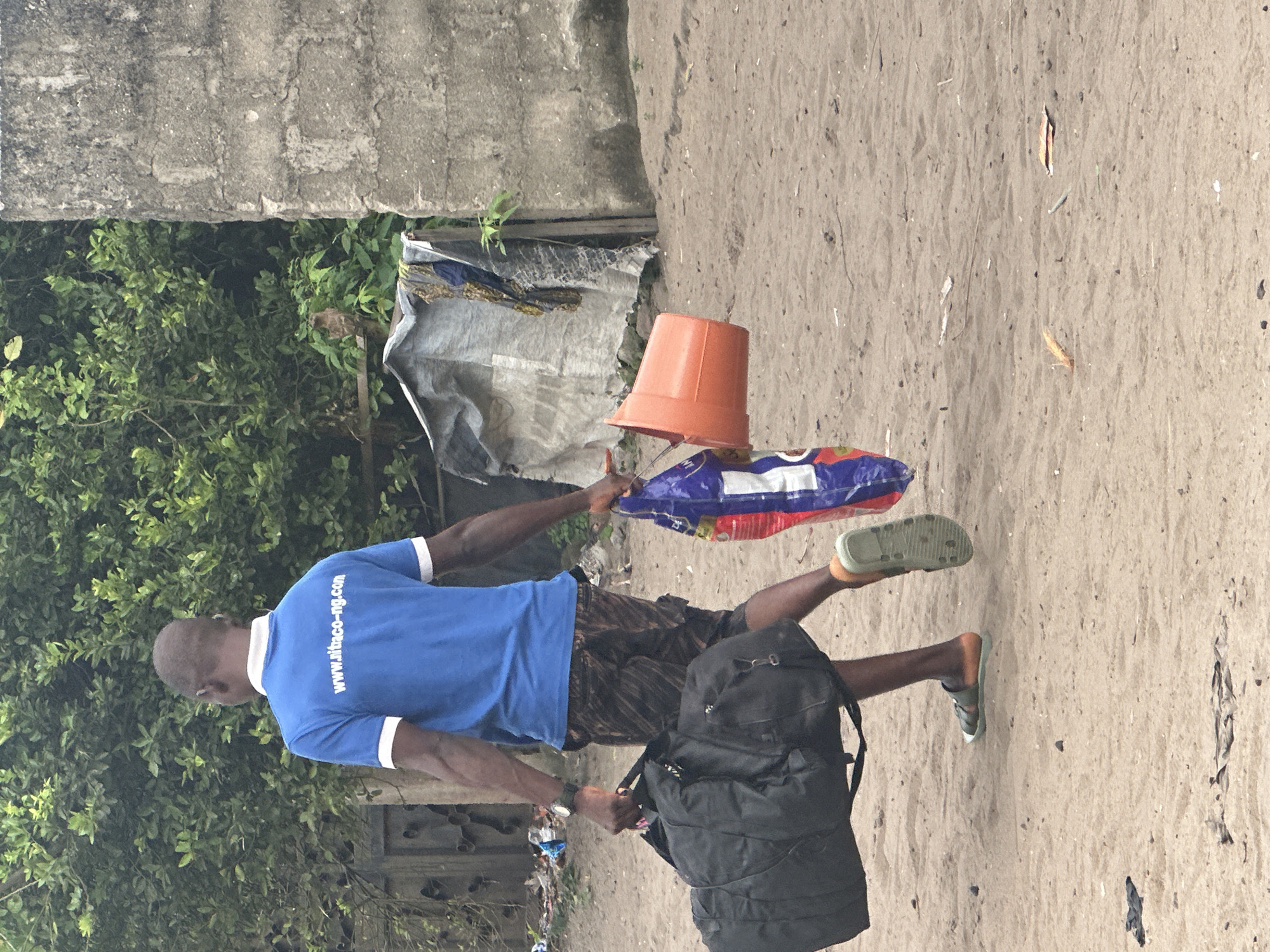 A Black man walks on a sandy path in Badagry, Nigeria, carrying a large black duffel bag in his left hand and an orange bucket and a blue and red plastic bag in his right. He wears a blue t-shirt with white trim and the text "www.sitaco-ng.com" printed on the back in white. He also wears patterned shorts and brown sandals. Behind him, a wall made of concrete blocks and a dense green bush are visible. The ground is sandy with scattered debris.