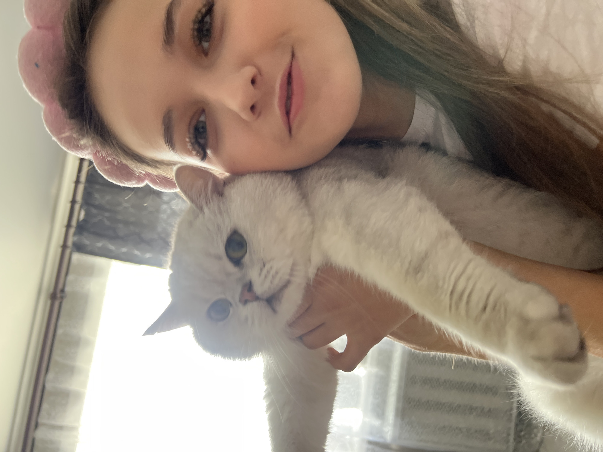A close-up shot features a fair-skinned young woman with light brown hair. She is wearing a pink headband and a white top. Her face is positioned close to a white cat, which is held in her hands. The cat has pale fur and visible green eyes. A window is visible in the background. The location is indeterminate.
