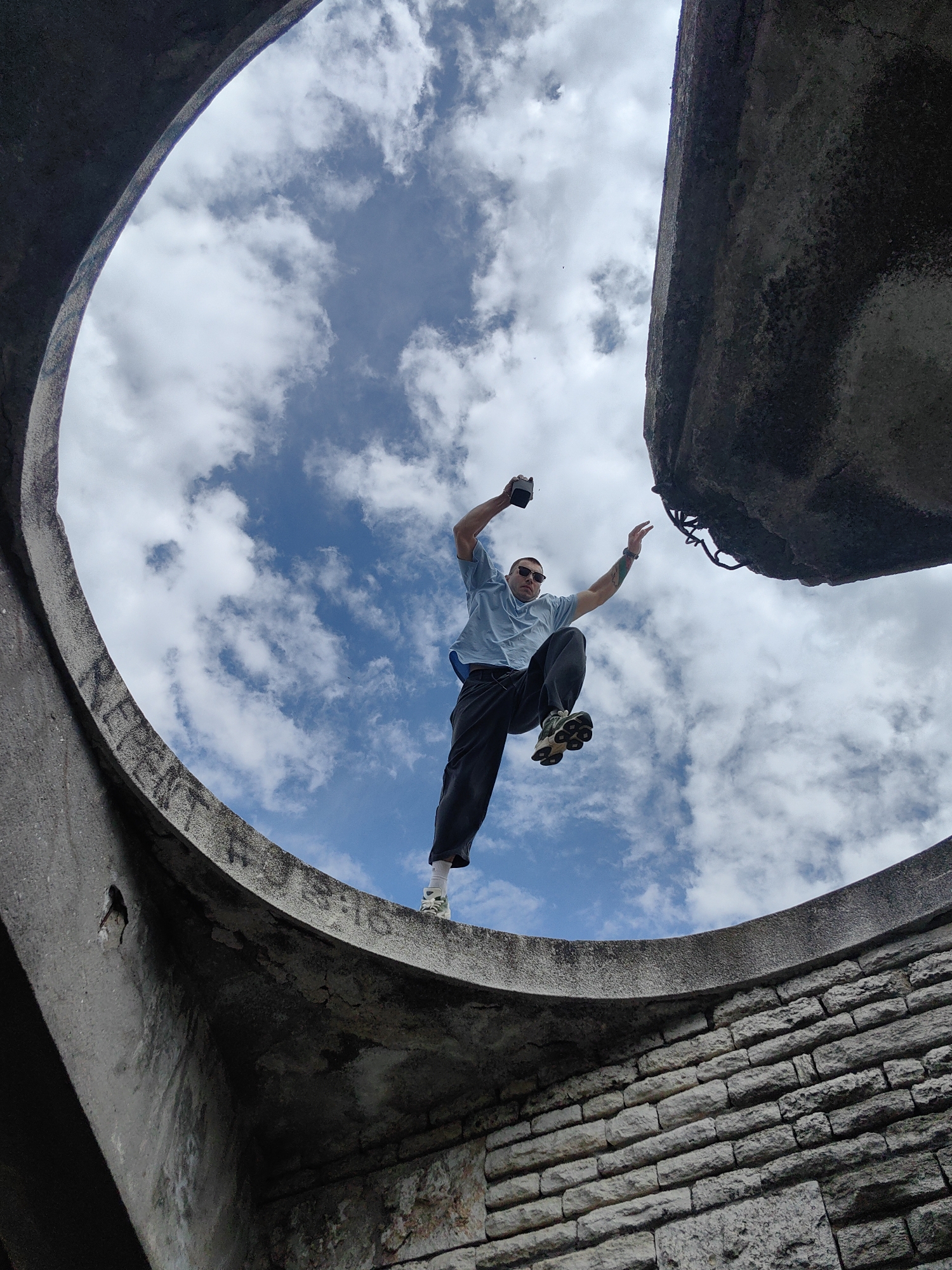 A man in sunglasses, a blue T-shirt, and black pants is leaping between two circular, concrete structures with a cloudy sky visible through the opening. He is holding a black object in his left hand, and his arms are outstretched. The structures appear to be ruins made of concrete and brick. This image likely depicts the Asparuhovo Bridge in Varna, Bulgaria.