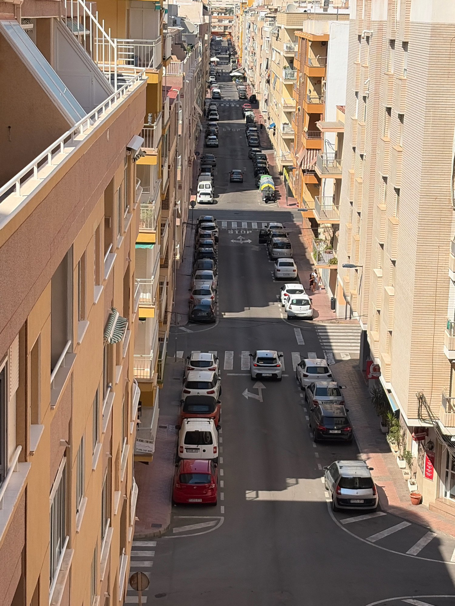 This is a high-angle, eye-level shot of a narrow, residential street in Torrevieja, Spain, likely during daylight hours given the strong shadows. The street is lined with multi-story apartment buildings on both sides, characterized by their beige and cream-colored facades, balconies, and numerous windows. Cars are parked along both sides of the street, creating a somewhat congested appearance.
On the left side of the street, the building is painted a warm, sandy orange and features white railings on its balconies and along an exterior staircase. The right side of the street is dominated by a pale, cream-colored brick building with dark-framed windows.
Several vehicles are visible, including sedans, a white van, and a small utility vehicle with a green tarp covering its load. Traffic markings, including white lane dividers and pedestrian crosswalks, are present on the asphalt road. Two individuals are standing on the sidewalk on the right side of the street, near a building entrance. The scene suggests a typical urban residential environment with limited space and reliance on street parking.