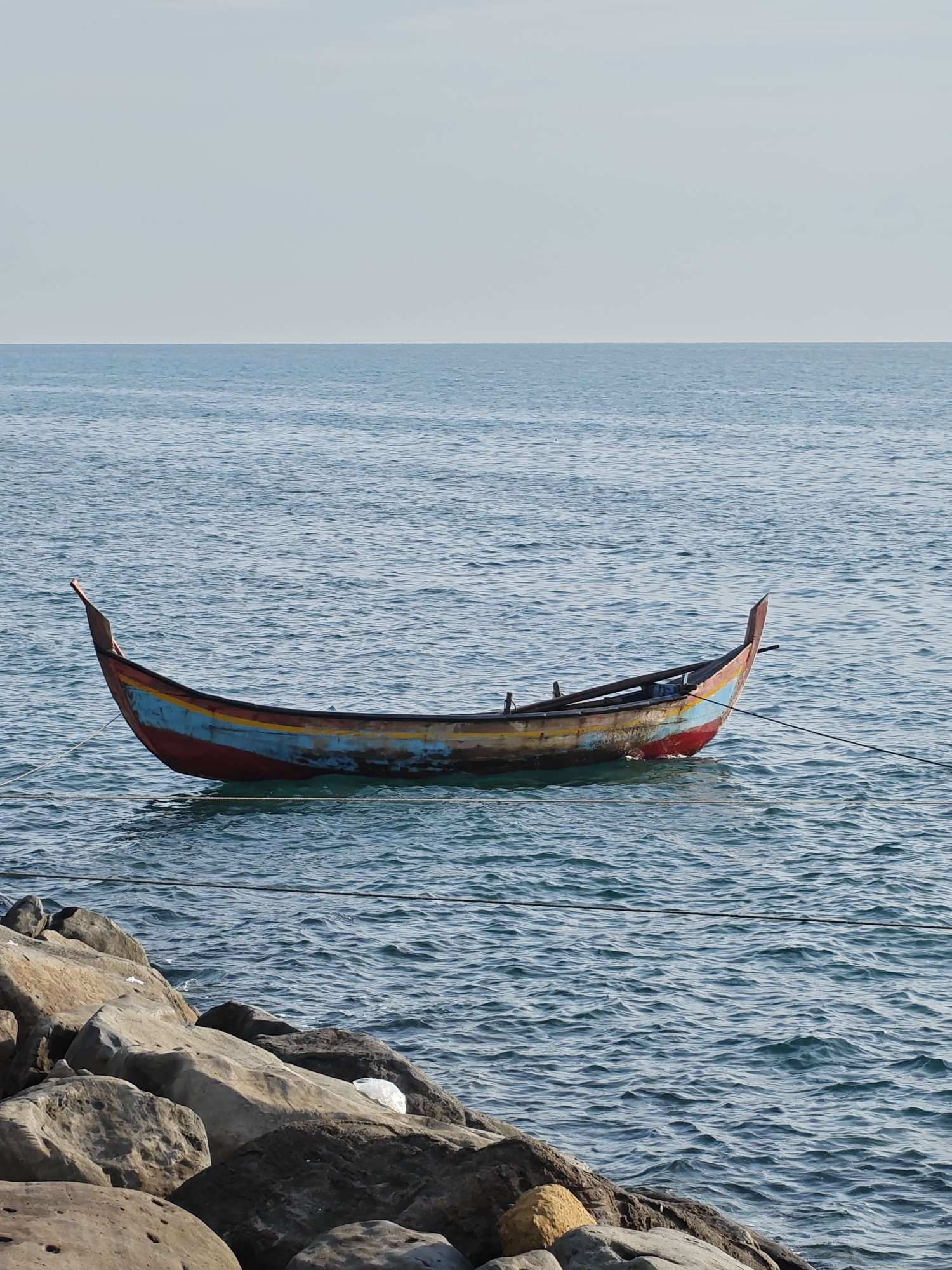 The image depicts a tranquil coastal scene in Lhokseumawe, Indonesia, centered around a traditional wooden fishing boat moored on calm waters. The boat, the main subject, is painted with vibrant, yet somewhat weathered, horizontal stripes of red, blue, and yellow on its hull, and features distinct, upward-curving ends. It appears to be secured by dark ropes, which stretch horizontally across the frame in front of it, indicating it's tethered to the shore or an anchor.
The sea exhibits a clear, deep blue-green hue, with gentle ripples visible across its surface, suggesting calm weather conditions. A clean, sharp horizon line separates the ocean from a pale, almost cloudless sky that occupies the upper portion of the image, appearing a uniform light blue or grey.
In the foreground, a rugged rocky shore or breakwater composed of large, irregularly shaped stones in shades of grey, brown, and ochre forms the bottom edge of the scene. A small white plastic bag can be seen resting on one of the rocks, a subtle detail of human impact. The lighting is bright and even, characteristic of daytime, likely midday or early afternoon, contributing to the peaceful and clear atmosphere of the scene. No people are visible, and there is no discernible text in the image.