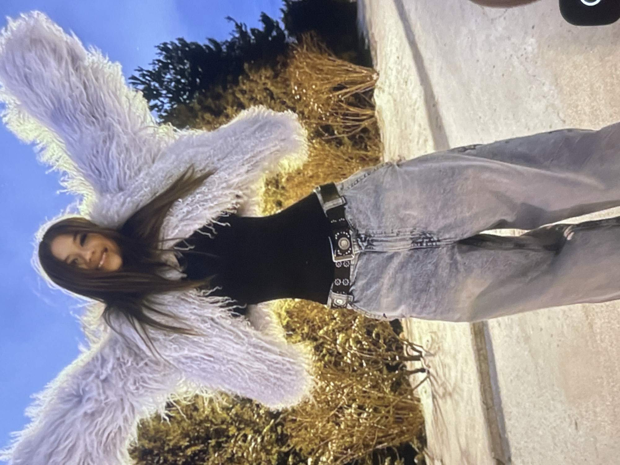 The image, captured in Poltava, Ukraine, features a young woman smiling brightly at the camera in an outdoor setting. She has long, dark brown hair and is wearing a distinctive, light-colored, fluffy or shaggy jacket that appears to be off-white or very pale grey. Beneath the jacket, she wears a fitted black top and light-wash, loose-fitting jeans. Her outfit is completed with a dark belt featuring a prominent, ornate silver-toned buckle.
The woman is standing with her arms outstretched, creating a "wing-like" effect with the voluminous sleeves of her jacket. The photograph is taken from a low angle, looking upwards at her, against a clear blue sky. In the background, various trees and shrubs are visible; some have dark green foliage, while others display dry, golden-brown branches or grasses, suggesting a natural environment. A light-colored, textured ground surface, possibly concrete or pavement, can be seen to her right. The overall mood conveyed is joyful and carefree. There is no visible text in the image.