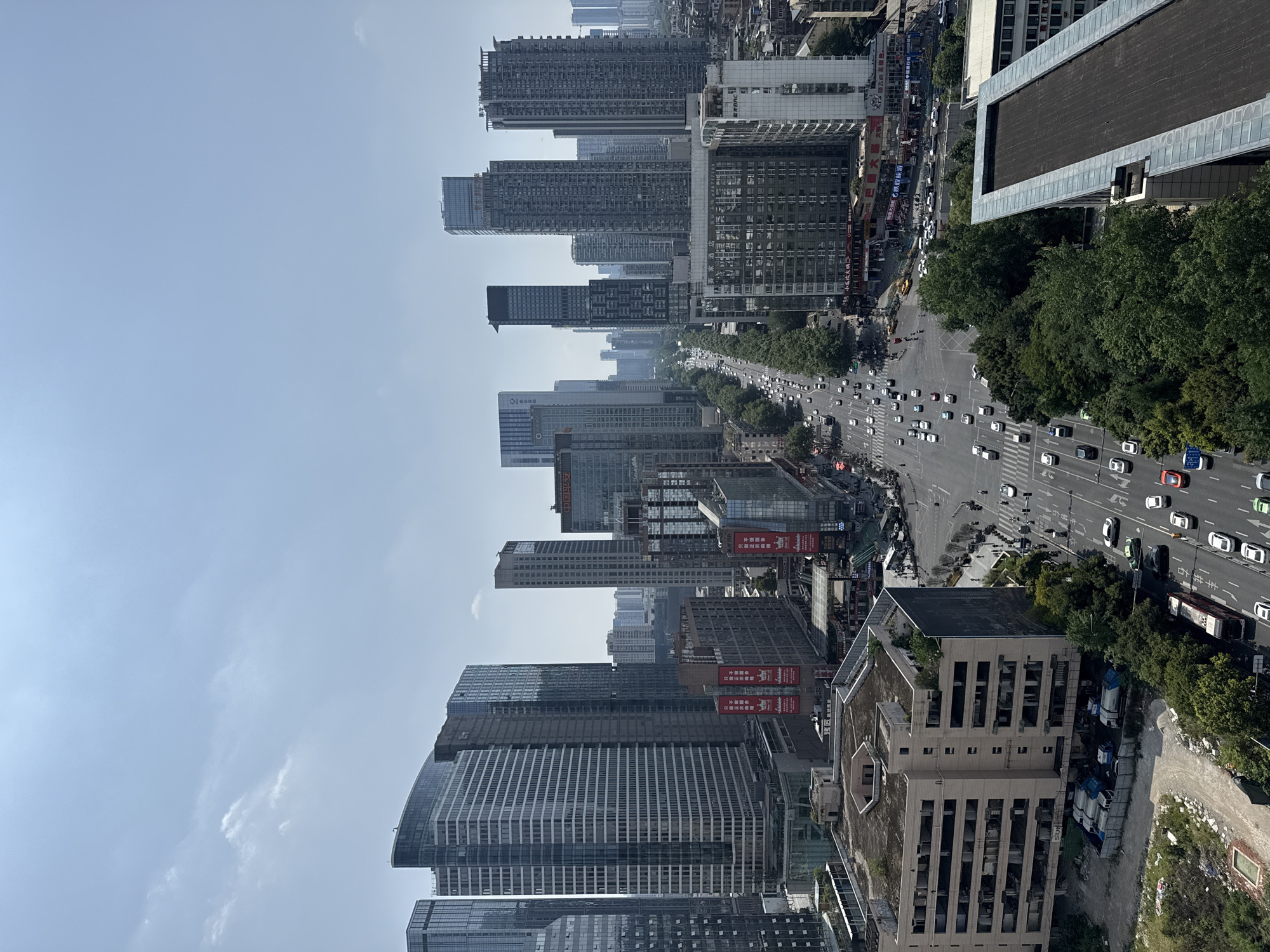 The image depicts a high-angle view of a street in Jinjiang District, Chengdu, China. The scene is dominated by a wide road filled with vehicle traffic moving in both directions. High-rise buildings line both sides of the street, suggesting a dense urban environment. There is some foliage lining the side of the road to the right, including trees. The sky is mostly clear with slight haze. The buildings feature modern architecture.