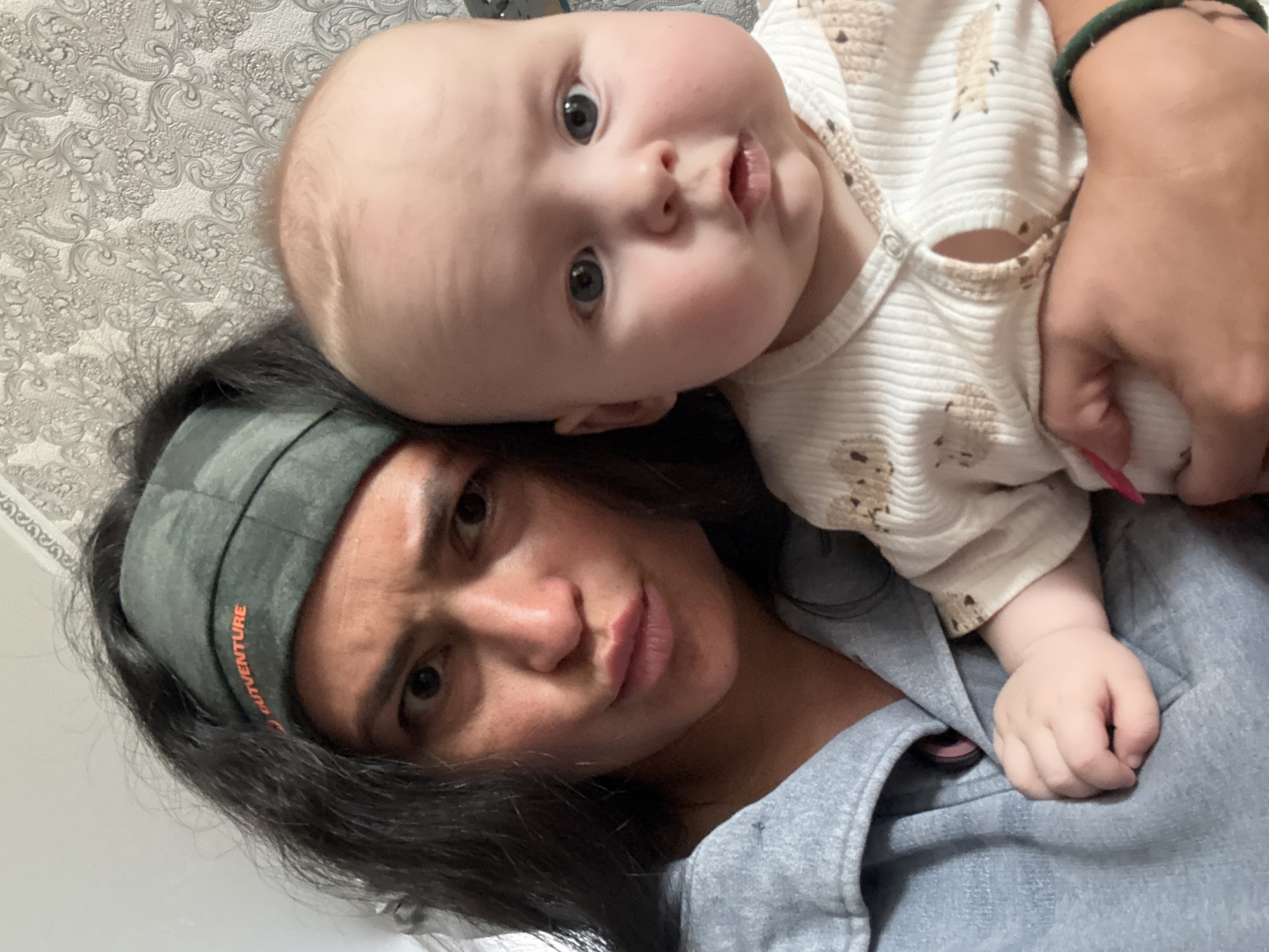 This intimate, tilted indoor photograph captures an adult woman with dark hair and a baby with light hair, appearing to be a mother and child. The woman, wearing a dark green headband and a grey top, has a soft, slightly pouting expression while looking directly at the camera. She also wears a dark green bracelet on her right wrist. The baby, nestled above and to the side of the woman's head, gazes with wide, curious eyes directly at the camera. The baby is dressed in a light-colored, long-sleeved onesie patterned with small, light brown figures, possibly hedgehogs or bears. The background features a light-toned wall covered in an intricate, decorative wallpaper pattern, suggesting a residential setting. Visible orange text on the woman's headband reads "OUTVENTURE." The overall impression is a personal moment captured in what appears to be a home in Кемуль, Russia.