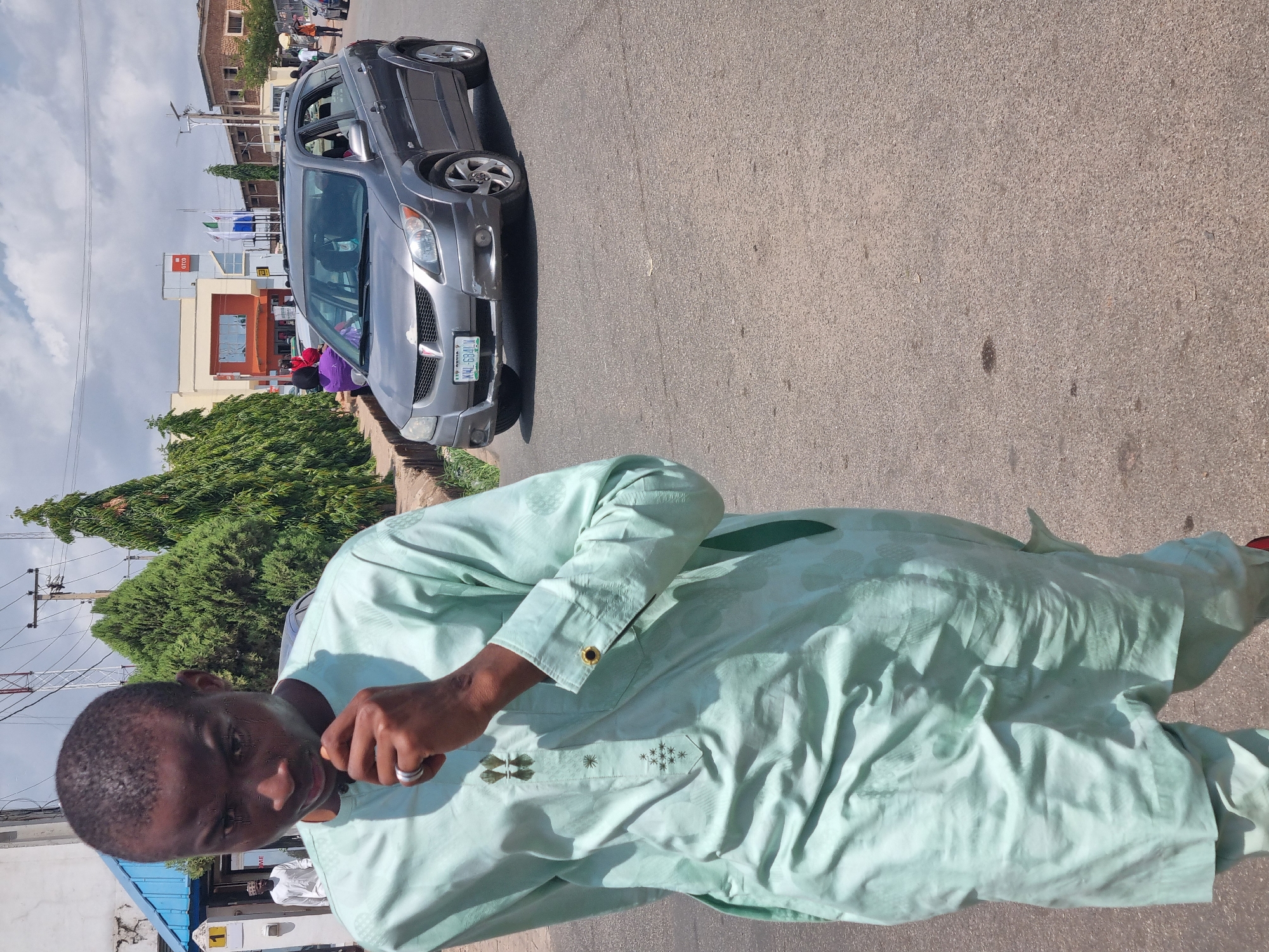 A man wearing a light green tunic stands on an asphalt road in Jos, Nigeria. Behind him, a silver Pontiac Vibe is parked on the side of the road. A building with a "GTCO" sign is visible in the background, along with trees and utility poles. People are walking in the distance near the building. The car has a license plate that reads "KWI-684LV".