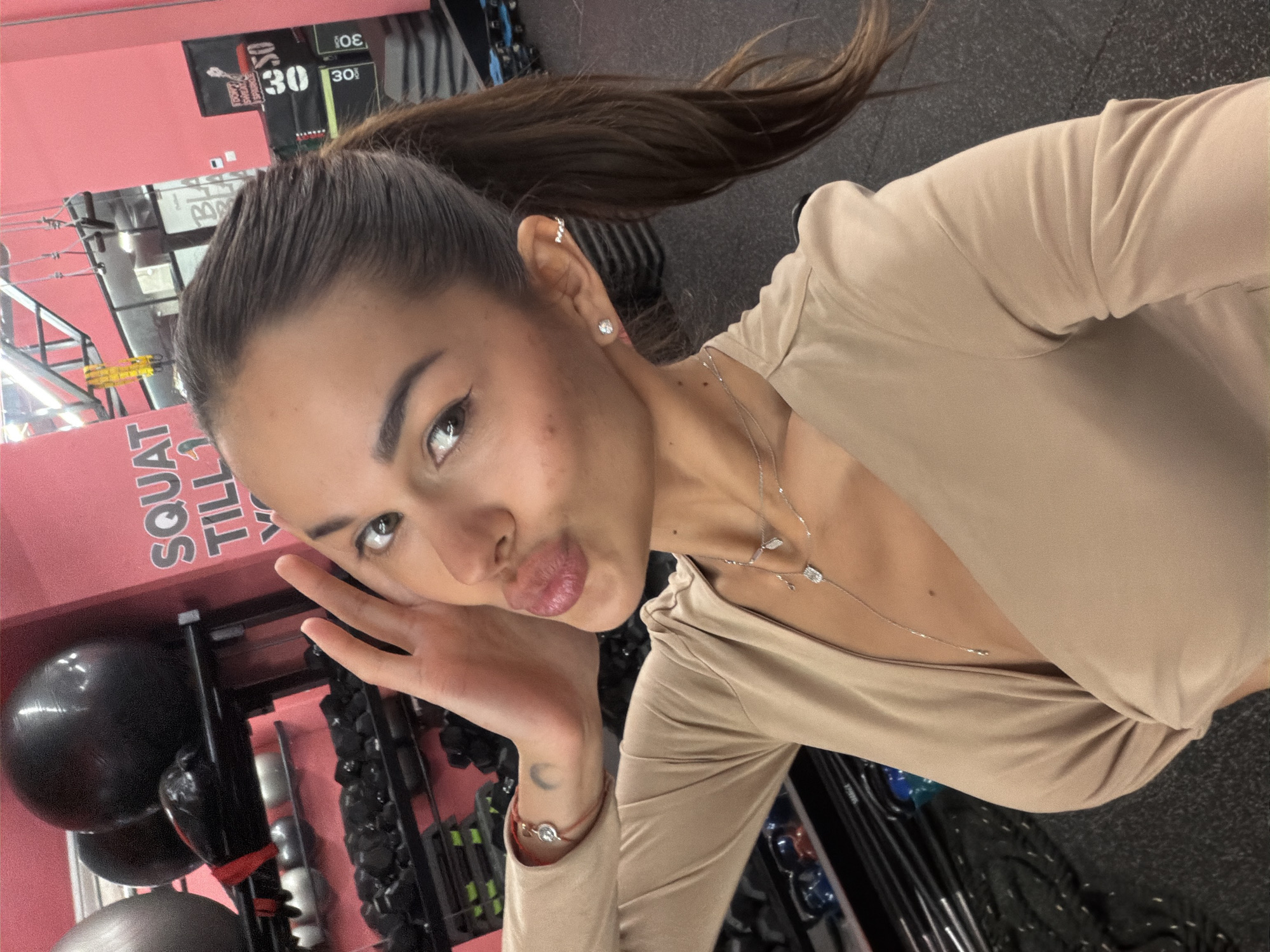 The image captures a female subject posing for a close-up selfie inside a fitness studio identified as Studio 30 in Tel-Aviv, Israel. The subject, an adult with long dark hair pulled back into a ponytail, presents with olive skin and light brown eyes, enhanced with visible eyeliner. She is wearing a light beige, long-sleeved top featuring a deep V-neckline and a gathered fabric detail at the torso. Accessories include a layered silver-toned necklace with multiple small pendants, including a rectangular diamond-like charm and a leaf-shaped charm, as well as diamond stud earrings and a silver ear cuff on her right ear. A red string bracelet with a silver charm is visible on her left wrist, below which a crescent moon tattoo is present on her forearm. Her lips are pursed in a 'kissing' expression, and her left hand is raised to her face, with fingers splayed near her cheek.
The background of the scene features distinct bright pink walls and black rubberized gym flooring. Metal shelving units are visible, holding various exercise equipment including large black stability balls on the top shelf and numerous black kettlebells and dumbbells on lower shelves. Several signs are mounted on the pink wall: white lettering on the left partially reads "SQUAT TILL 1". To the right, a prominent black banner or box with white text includes phrases like "DON'T BE AFRAID OF FAILURE" (partially visible) and multiple repetitions of the number "30", confirming the gym's name, "Studio 30". Overhead, what appears to be suspension training ropes are visible hanging from the ceiling structure. A reflective surface, likely a window or glass partition, is present in the upper central background, showing indistinct reflections.