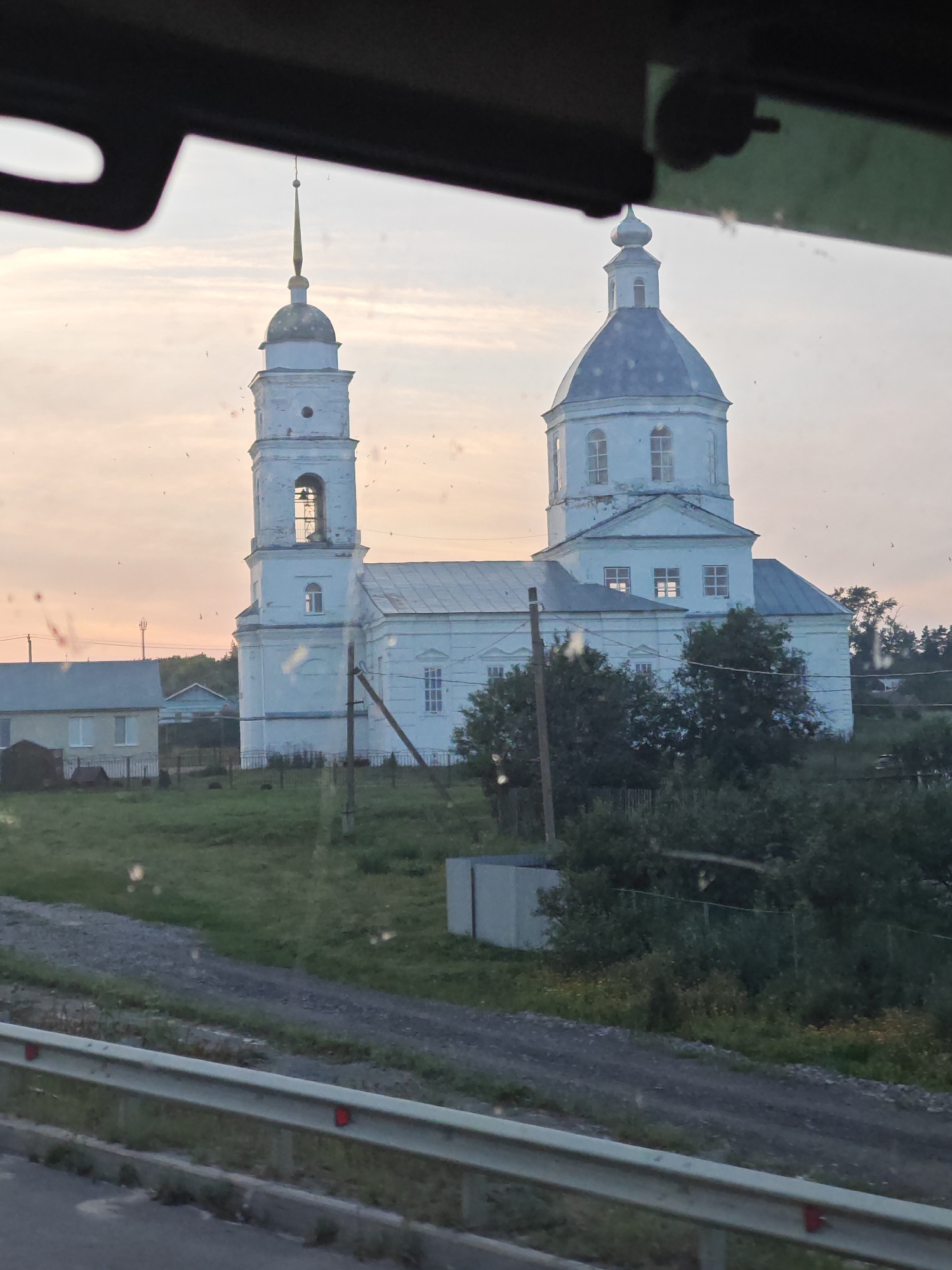 The image depicts a white church with a tall bell tower and a dome, situated in a grassy, rural landscape. The sky is overcast with a pale orange hue, suggesting either sunrise or sunset. In the background, there are a few simple buildings, a gravel road, and dense trees. The foreground shows a metal guardrail, indicating the photograph was taken from a moving vehicle. The church appears to be the central focus, with its distinct architectural features highlighted against the soft lighting of the sky. There are no people or animals visible in the scene. The overall atmosphere is tranquil and somewhat muted due to the lighting conditions. The location is identified as the city of Симбухово, Russia.