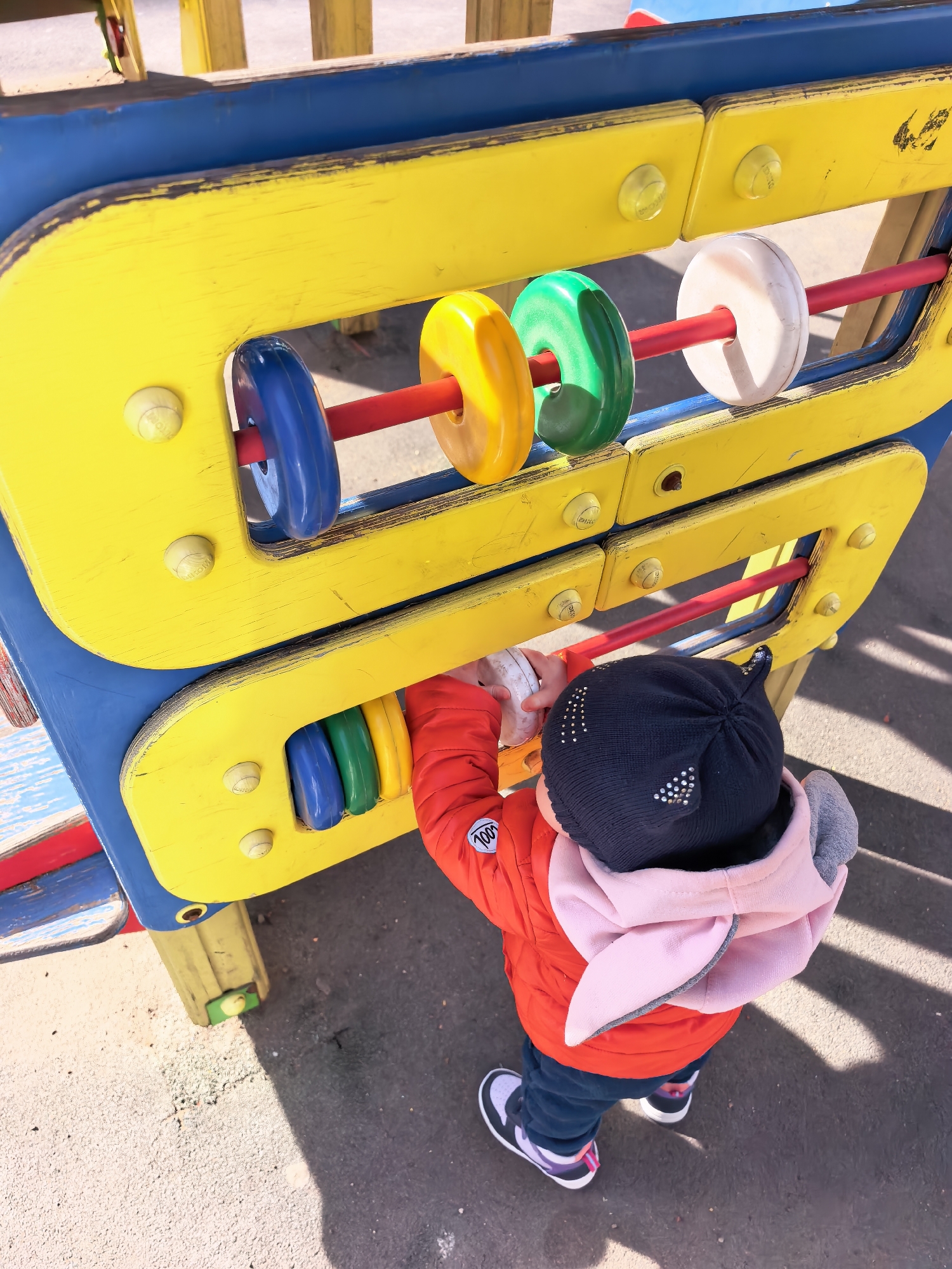 A young child in a red jacket, dark pants, and black-and-white sneakers is playing with an abacus-like toy on a playground. The toy consists of two rows of colorful, circular beads strung on red rods within a yellow and blue frame. The child, wearing a black beanie with silver embellishments and a pink hooded jacket, is seen from behind, reaching for the beads on the top row. The playground appears to be located outdoors on asphalt. Based on available information, the playground may be located in Sterlitamak, Russia.