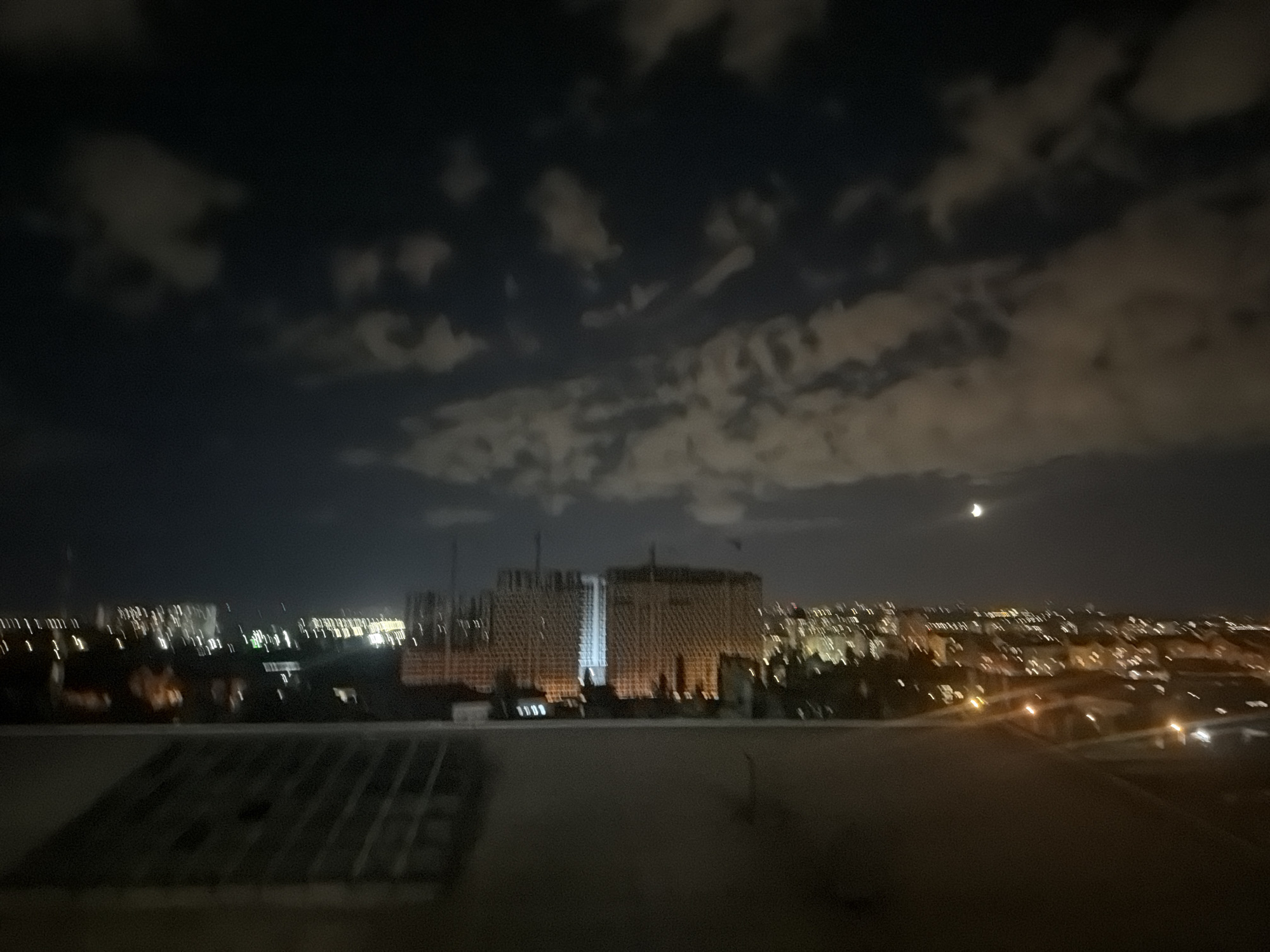 This is a blurry, nighttime photograph of an urban landscape. In the foreground, there is a dark, elevated surface, possibly a rooftop, with a faint, grid-like pattern visible. In the middle ground, a large, illuminated building under construction dominates the scene. It appears to be a multi-story structure with a complex facade and scaffolding. The building is brightly lit, with sections of its exterior glowing white. Beyond this construction site, the city lights of Kyiv, Ukraine, are visible stretching into the distance. The lights are blurred and create streaks of yellow and white against the dark sky. The sky itself is dark, with patches of clouds illuminated by ambient light. A crescent moon is visible in the upper right quadrant of the image. The overall impression is of a developing or bustling urban area at night.