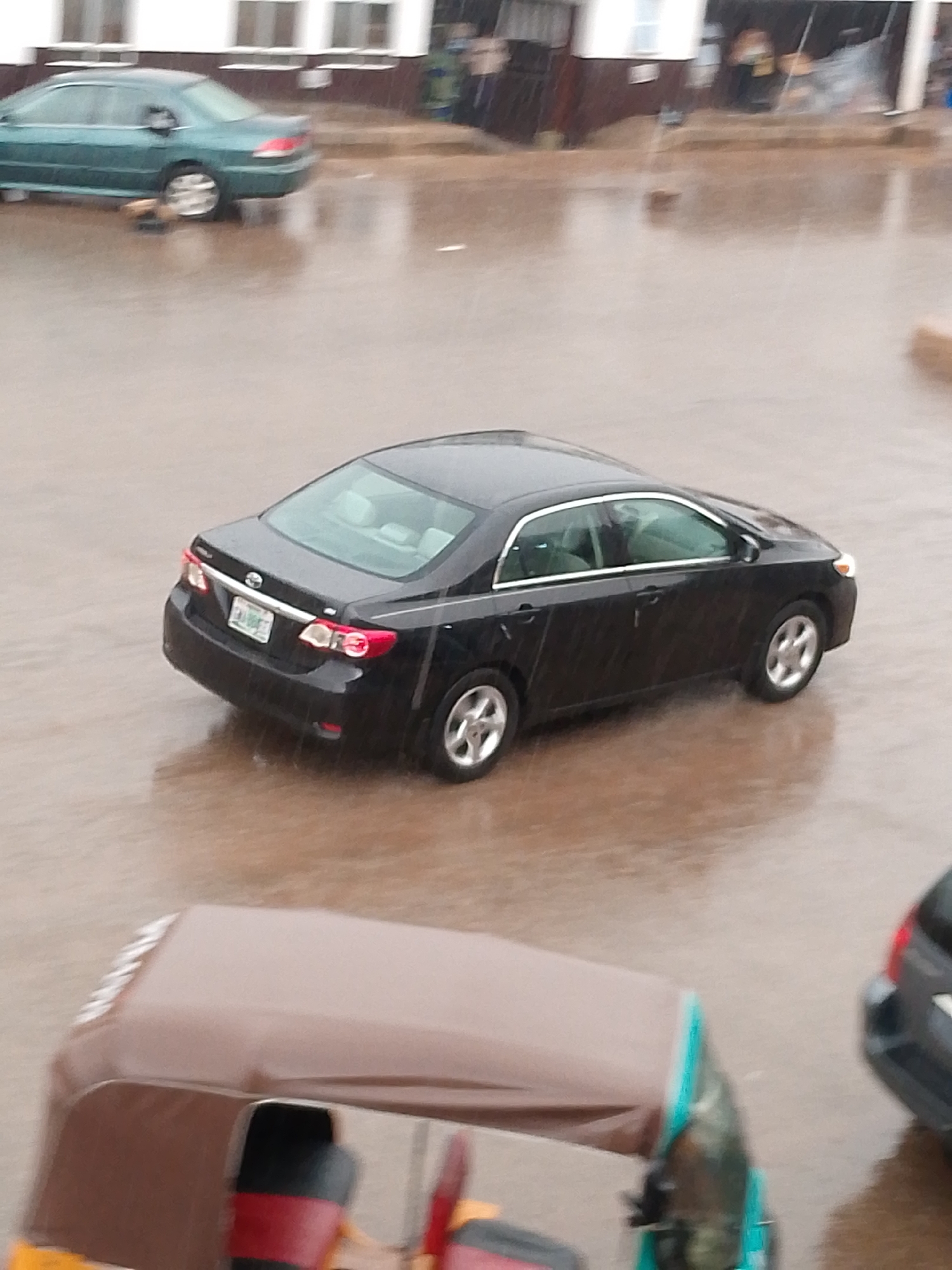 This media file depicts a street scene in what appears to be Bukuru, Nigeria, during a heavy rainfall. The ground is wet and reflective, indicating the presence of puddles and standing water, suggesting a flooded or waterlogged environment.
The main subject is a black sedan, likely a Toyota Corolla, seen from a rear three-quarter view. It is parked or moving slowly on the wet road. The car's taillights and license plate are visible, though the text on the license plate is difficult to decipher due to the image quality and distance. Another car, a green sedan, is visible in the background on the left side of the frame, partially obscured by the foreground. In the foreground, a portion of a three-wheeled vehicle, possibly an auto-rickshaw or tuk-tuk, is visible. Its roof is a tan color, and parts of its seating appear to be red and black.
The weather is clearly rainy, with visible streaks of rain in the air and on the surfaces. This suggests a gloomy or overcast day. The setting appears to be an urban or semi-urban street with buildings visible in the background. The overall atmosphere is one of a wet, possibly inconvenient, day due to the downpour. There are no discernible people actively engaged in the scene, but the presence of vehicles suggests ongoing activity despite the weather. The time of day is not explicitly indicated, but the overcast conditions associated with heavy rain could suggest it is daytime, perhaps late morning or afternoon.
