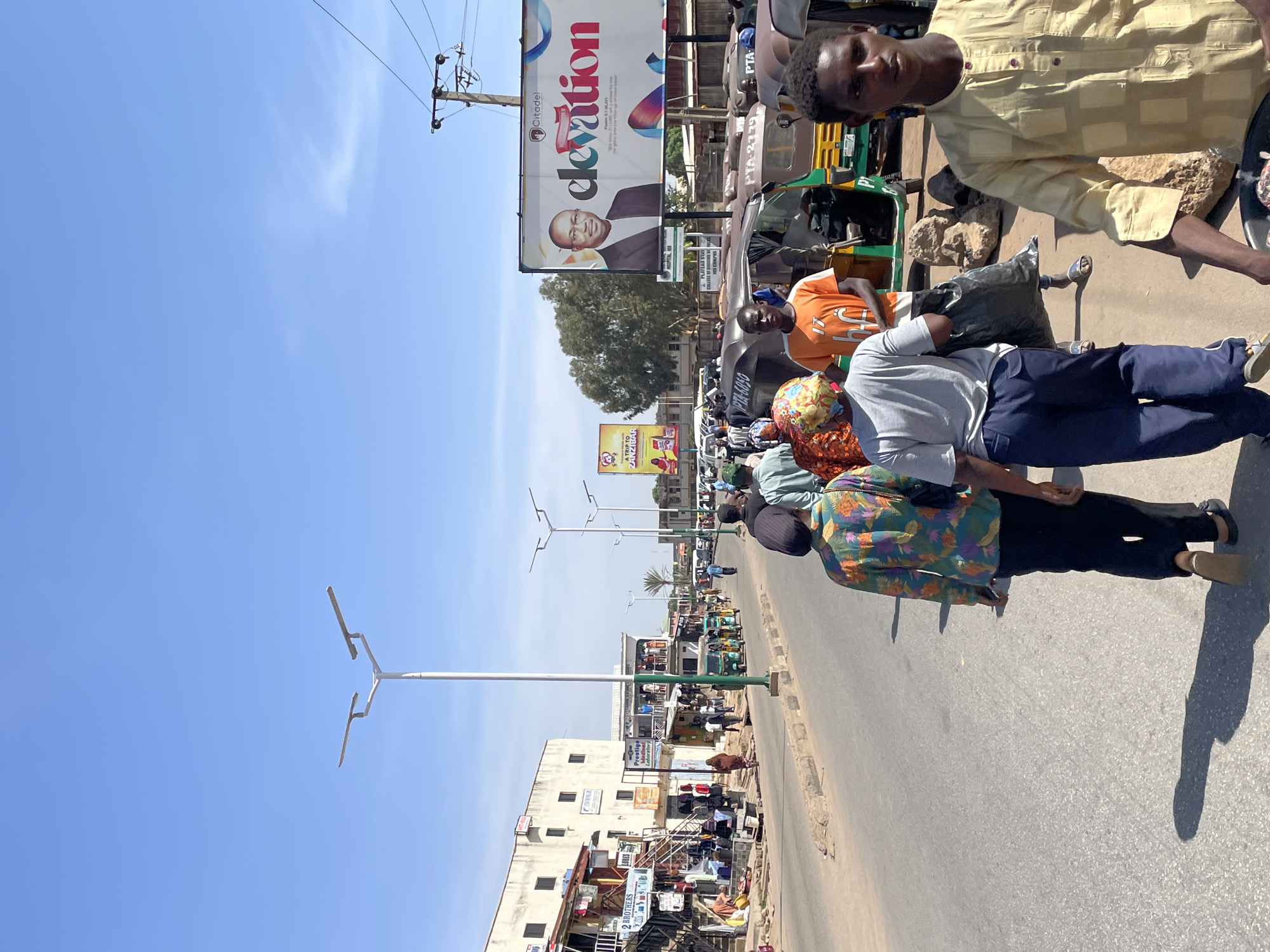 This media file captures a vibrant street scene in Jos, Nigeria, under a bright, clear blue sky during the daytime, indicated by the strong sunlight and distinct shadows. The image portrays a bustling urban environment with a mix of people, vehicles, and commercial buildings.
Several individuals are visible on the asphalt road and sidewalks, engaged in daily activities. In the immediate foreground, a young man wearing a light yellow patterned shirt looks directly at the camera. Other people, dressed in casual attire including a grey t-shirt and blue pants, and an orange t-shirt, are walking or standing, some appearing to be commuters or involved in street-side commerce.
The road is populated by numerous tricycle taxis (Keke Napep), some parked and others seemingly in motion. These vehicles display partial registration numbers such as "PTA-2779" and "PTA-6850." The street is lined with multi-story commercial buildings, featuring storefronts and signs. Visible text on these establishments includes "2 BROTHERS," "Prestige Laboratory," "PLATEAU'S," "RES," and "HOSPITAL NS."
Overhead, utility poles with power lines and modern streetlights define the infrastructure. A prominent large billboard in the mid-ground advertises "Citadel" and "elevation," featuring a man in a suit and the slogan, "We Help Small and Medium Businesses to Grow their profits and stand them tall in the market." Further down the street, a smaller billboard for "ZANZIBAR" is also visible. The scene conveys a typical busy day in a Nigerian city.