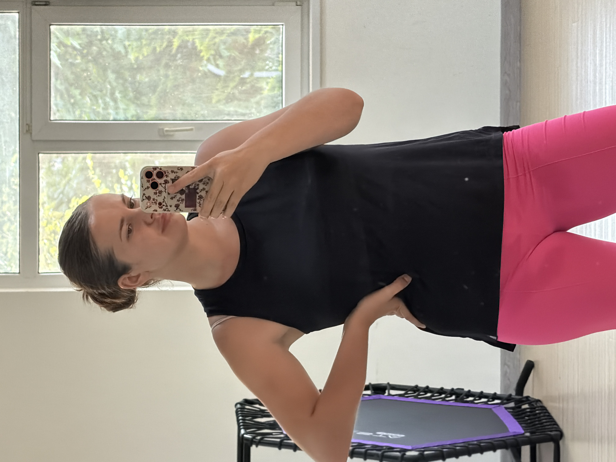 A young woman with her hair pulled back in a bun, wearing a black tank top and bright pink leggings, stands in a room with a trampoline to her left and a window in the background. She is holding a floral-patterned smartphone in her right hand, taking a selfie in a mirror. The room appears to be a gymnasium or fitness studio, as indicated by the presence of the trampoline and what appears to be a wooden floor. The window is large and reveals greenery outside, suggesting a sunny day. The woman is looking at her phone as she takes the picture. The image does not provide information to determine the specific location within the city of Sochi, Russia.