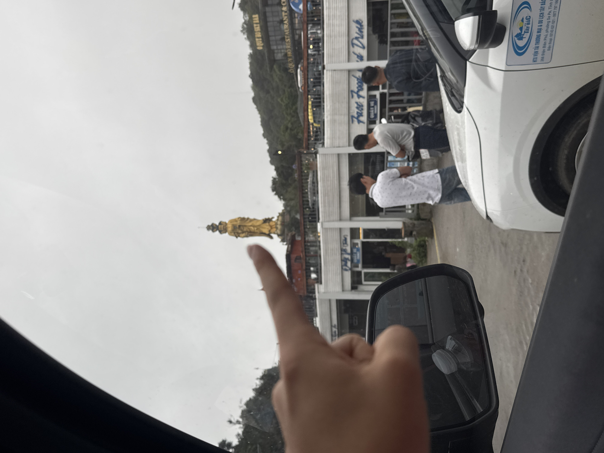 The image shows a view from inside a vehicle in Binh Lu, Vietnam. A person's hand is pointing towards a golden Buddha statue in the distance. Several buildings are visible, including "O QUY HO SKY BAR" and "Fast Food & Drink" establishments. A group of three men is standing nearby. A white car with "HTX VẬN TẢI THƯƠNG MẠI & DU LỊCH TÂY BẮC" printed on the side is parked next to the vehicle. The weather appears overcast. The car's side mirror is visible.