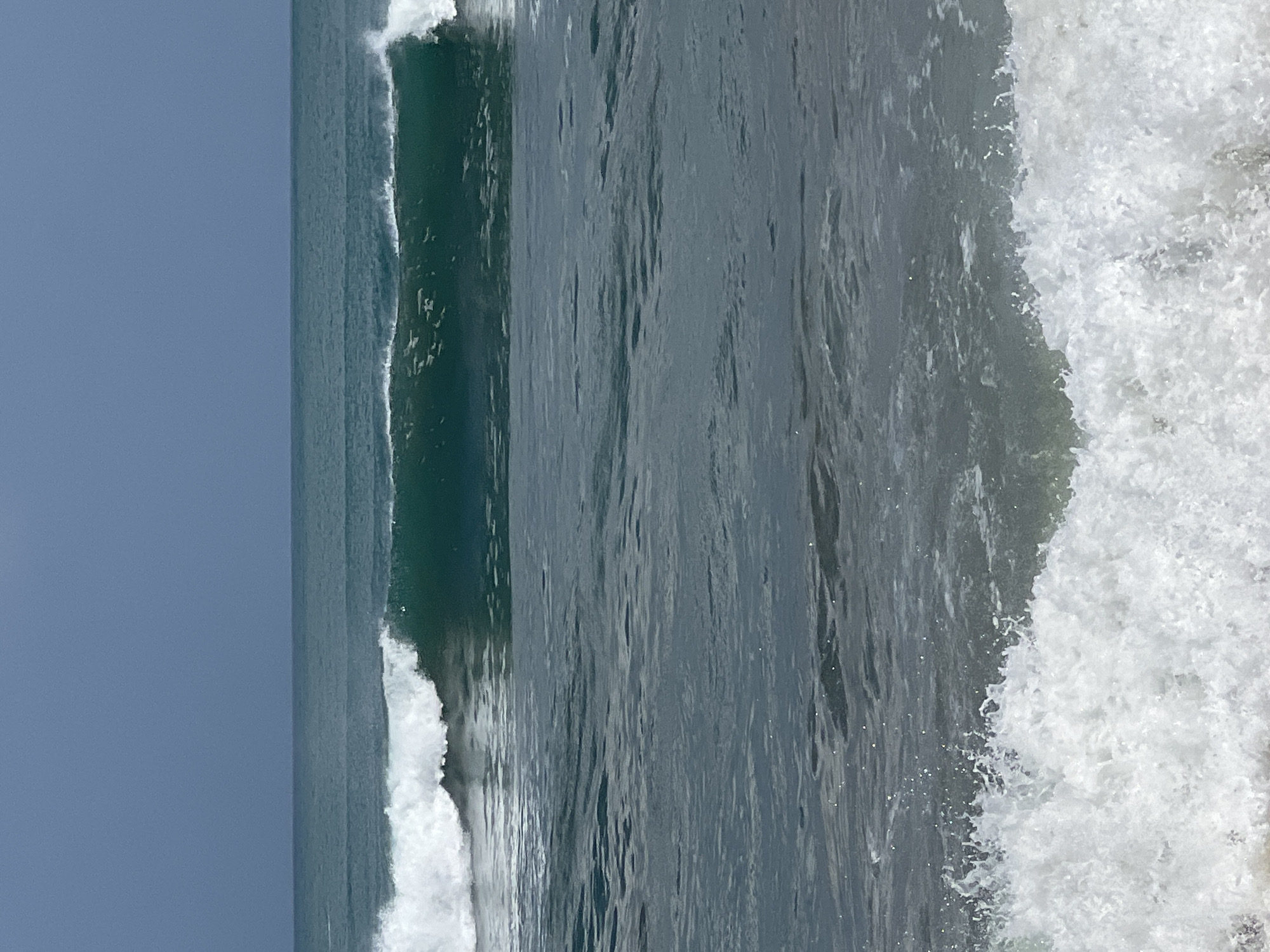 The image displays a serene ocean scene with a clear blue sky. The ocean is calm, with gentle waves rolling towards the shore. In the mid-ground, a larger wave is breaking, creating a translucent green crest with white foam. The foreground shows the foamy churn of waves nearing the beach. The horizon line is distinct, separating the deep blue sky from the teal-colored ocean. There are no people or structures visible in the frame. The scene captures the natural beauty of the ocean, likely taken from the coast of Kuta, Indonesia, a popular beach destination known for its surf and scenic views.