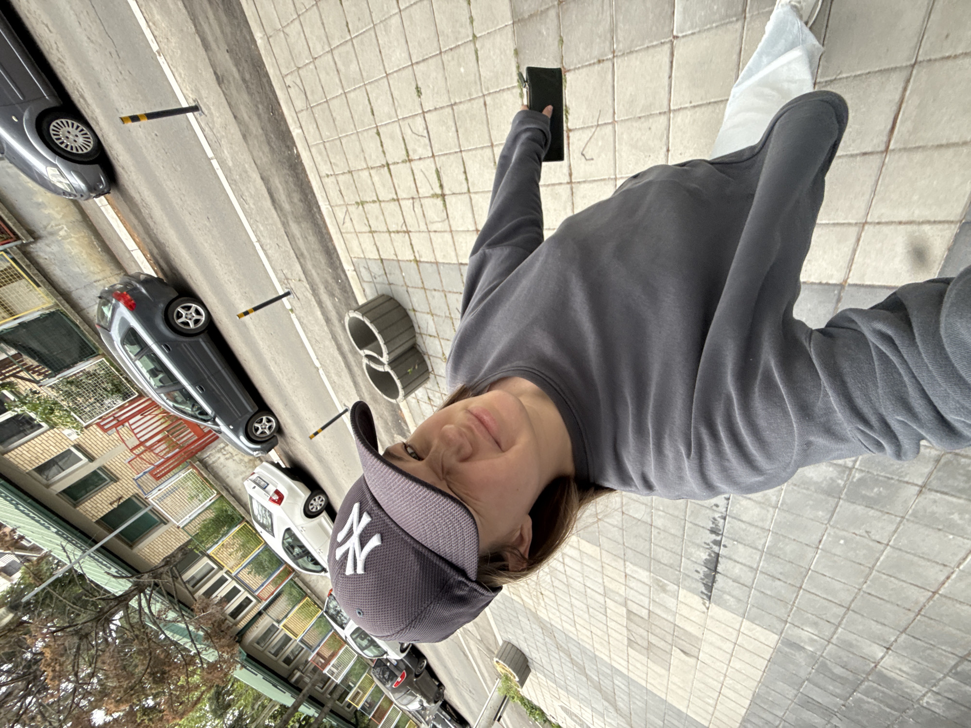 A person wearing a gray sweatshirt and a New York Yankees baseball cap is taking a selfie. The person is standing on a sidewalk made of square tiles. In the background, there is a street with cars parked along it and buildings with windows, some of which are covered with orange metal grids. The buildings appear to be located in the Vozdovac Urban Municipality, Serbia. The person is holding a black rectangular object, possibly a phone, in their left hand.