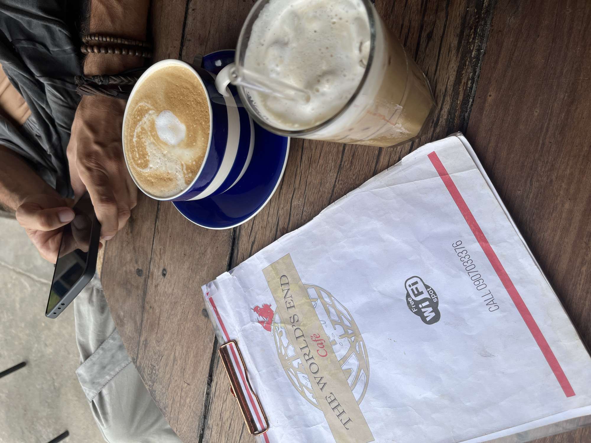 A top-down view captures a wooden table at "The World's End Cafe" in Koh Phangan, Thailand, where a person is using a smartphone. The table is set with a blue and white striped mug of latte art coffee, a glass of iced coffee with a straw, and a clipboard holding a menu. The menu features a globe graphic and the cafe's name, along with "Free Wi-Fi spot" and a contact number. The person's hands, adorned with beaded bracelets, are visible as they hold the phone. The environment suggests a casual outdoor setting with a concrete floor visible in the background.