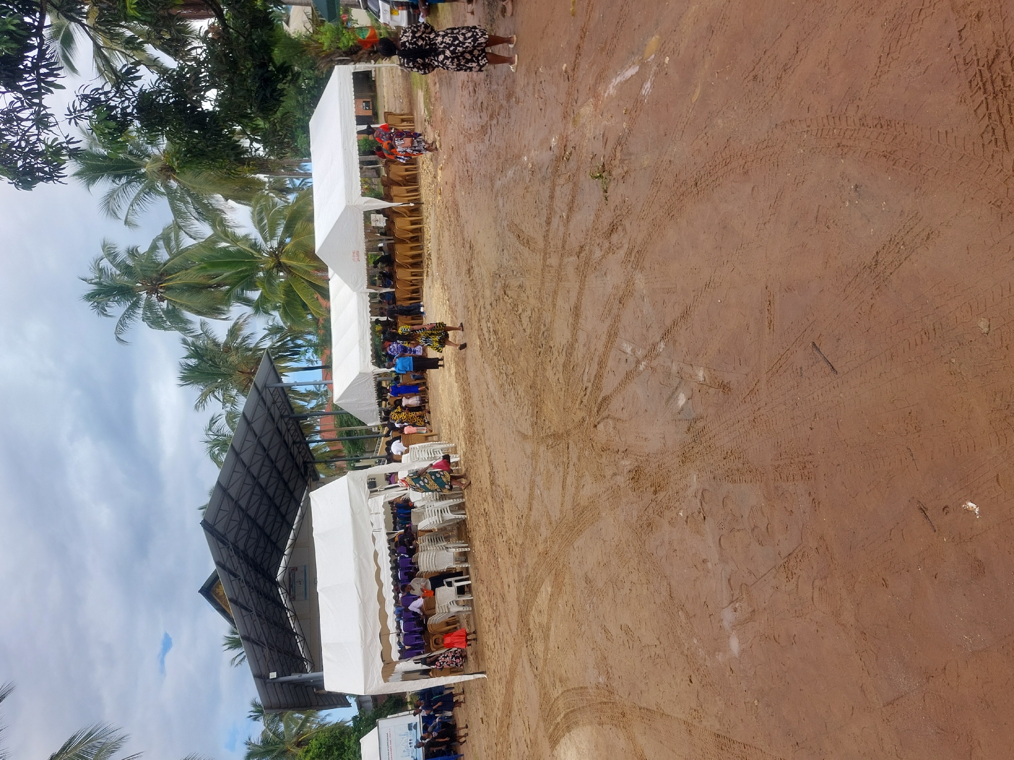 The image depicts an outdoor event occurring on a dirt or sand-covered ground in Dar es-Salaam, Tanzania. Several white tents are set up, with rows of chairs beneath each, occupied by attendees, predominantly dressed in colorful clothing. A stage with a dark-colored roofing structure is also present. Palm trees and other greenery are visible. There are tire tracks. People can be seen walking around the tents. The sky shows a mix of clouds and clear areas.