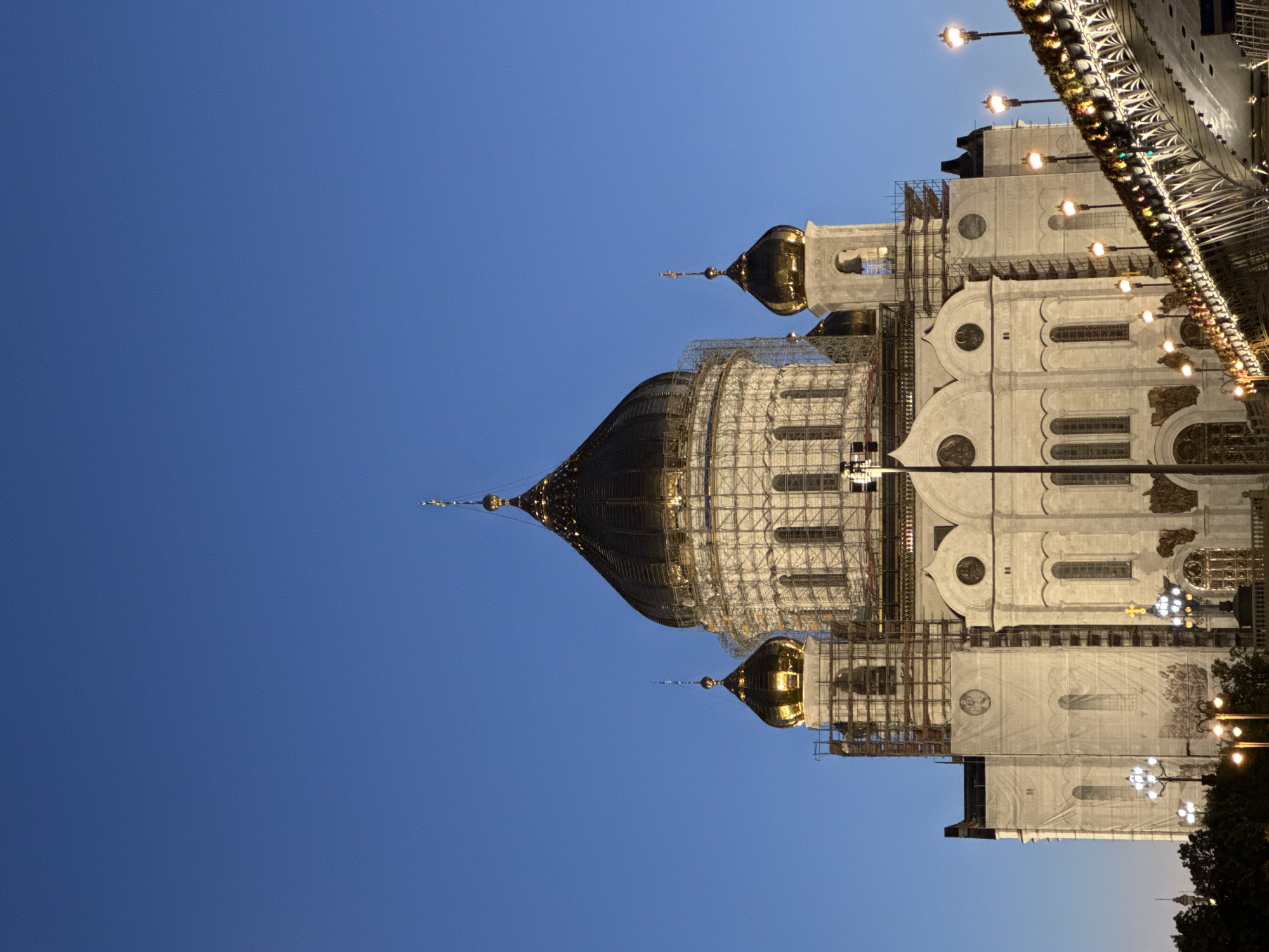 The image shows the Christ the Savior Cathedral in Moscow, Russia. The cathedral, a prominent landmark, is illuminated against a clear, deep blue twilight sky. Scaffolding partially covers the white stone facade and the large central golden dome, suggesting ongoing renovation or construction work. Smaller golden domes, topped with crosses, are also visible. A bridge adorned with festive lights and greenery spans the foreground, indicating the presence of a public space or thoroughfare. Several street lamps with warm lights are scattered throughout the scene, illuminating the area around the cathedral and the bridge. The overall atmosphere is serene, with the lights creating a striking contrast against the fading daylight and the architectural grandeur of the cathedral. There are no people visible in the frame.