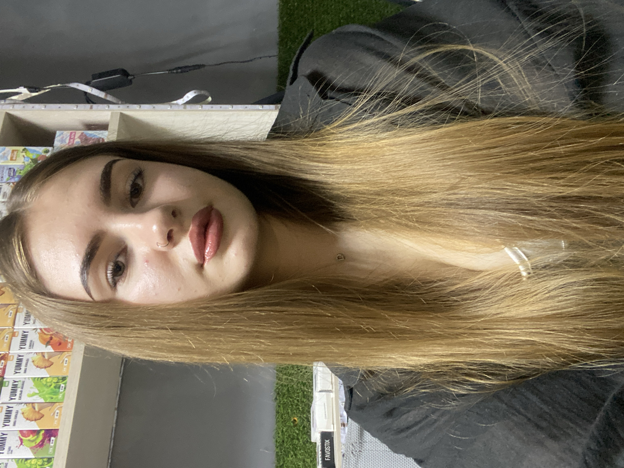 A medium shot photo shows a young woman with long, straight, honey-blonde hair. She has fair skin, full lips with a subtle sheen of lipstick, a small nose ring, and dark eyebrows. She is wearing a black collared jacket over a white top. A small silver necklace with a "D" pendant is visible. She is looking directly at the camera. The background features shelves stocked with various colorful boxes, a patch of artificial green turf, and some lighting elements. The image appears to be taken indoors. No information about the location in Дьяково, Russia is present in the image itself.