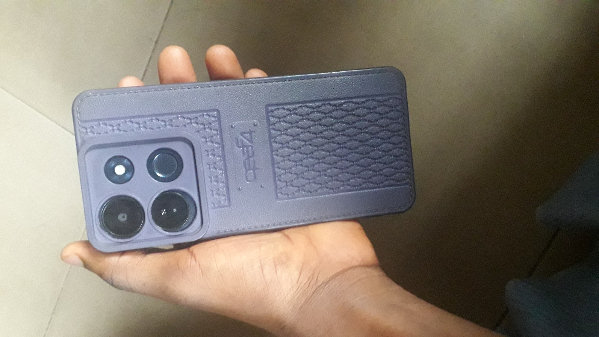 A person's hand is holding a dark purple smartphone case. The case has a textured pattern on the back, with a prominent camera module that houses four circular lenses arranged in a square pattern. The brand name "QEEA4" is visible on the case. The background suggests an indoor setting with a tiled floor. The image does not provide any specific landmarks or details to pinpoint a location within Nkpor, Nigeria.