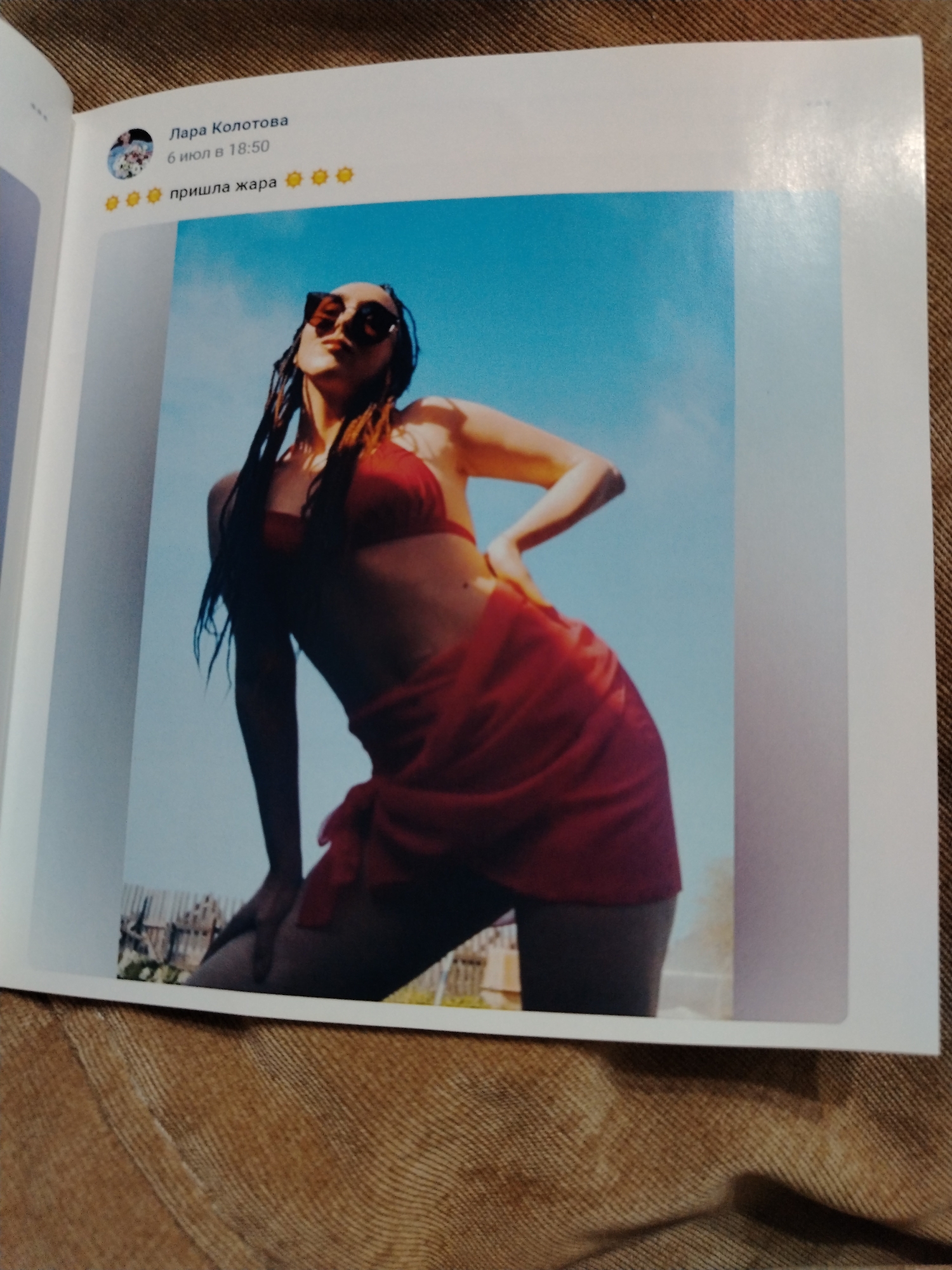 A photograph depicts a page from a book or magazine displaying a simulated social media post featuring a woman posing in a red bikini and sarong against a blue sky backdrop. The post, attributed to "Lapa Konotoba," includes the date "6 mon B 18:50" and the text "npuwna xapa." The woman is wearing sunglasses and has wet hair. The background of the photograph shows a blurry landscape. The photo is taken at a close, slightly elevated angle, and is located in Izhevsk, Russia.