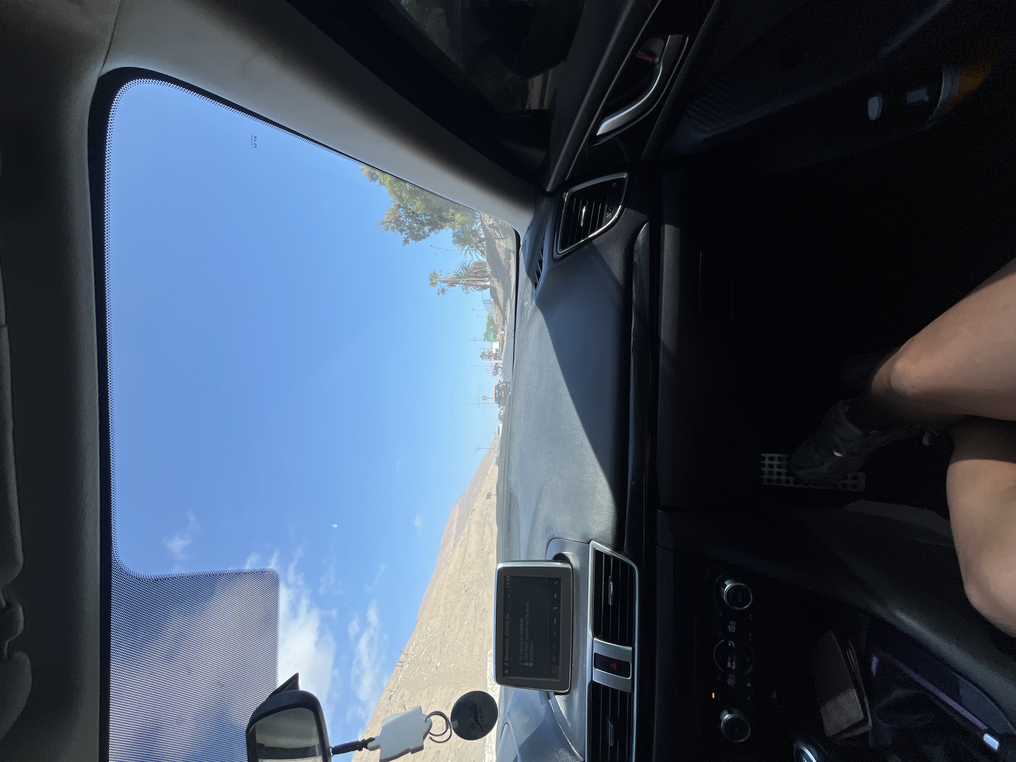 The image depicts a view from inside a car, looking out through the windshield. The outside scene includes a clear, blue sky with scattered clouds, a road leading towards a town with palm trees and buildings. A mountain is visible in the distance. Inside the car, the dashboard, steering wheel, and a display screen are visible. The driver's leg is also visible. The city location is unidentified.