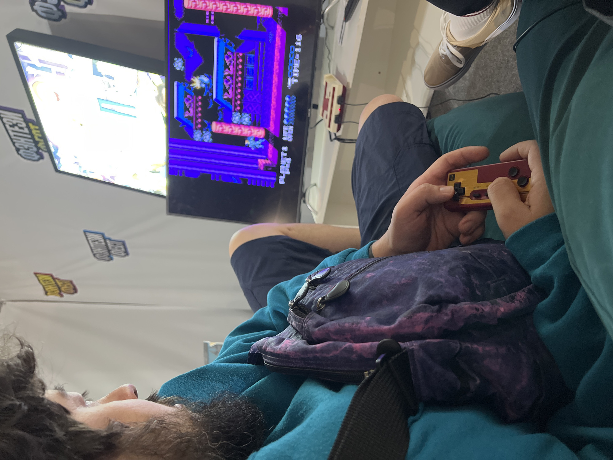 A man, facing left in profile, is playing a video game. He has curly dark hair and a beard. He wears a teal hooded sweatshirt and dark shorts. A purple and pink patterned bag is slung across his chest. He holds a red and yellow retro video game controller in his hands. The video game is displayed on a large television screen, showing a colorful scene with what appears to be a character on a platform. A retro video game console is positioned on a white shelf beneath the television. Two signs with Cyrillic text are visible on a white wall in the background. The man's feet are visible in the lower right corner; he wears brown sneakers and white socks. The scene appears to be indoors, possibly at an event or convention.