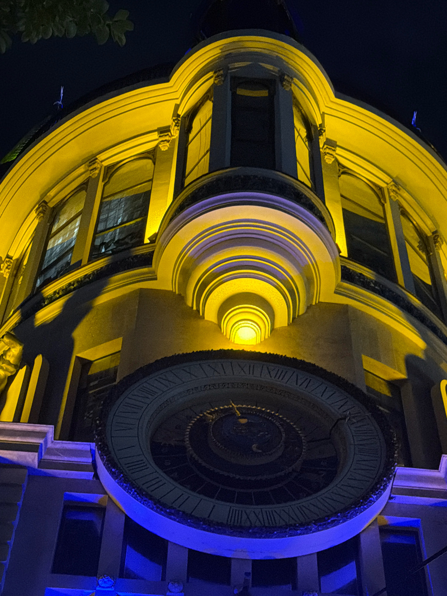 This is a low-angle, night-time photograph of a building, likely the Batumi Astronomical Clock, located in Batumi, Georgia. The building's facade is illuminated with warm yellow light, highlighting architectural details such as rounded cornices and arched windows with stained glass. A prominent circular structure, the clock face, is visible in the lower portion of the image, featuring Roman numerals and intricate designs. The clock face is framed by a blue light. Shadows are cast on the building, suggesting the presence of external light sources. A few silhouetted leaves from an overhead tree are visible in the upper left corner against a dark sky. There are no people or discernible interactions depicted in the image.