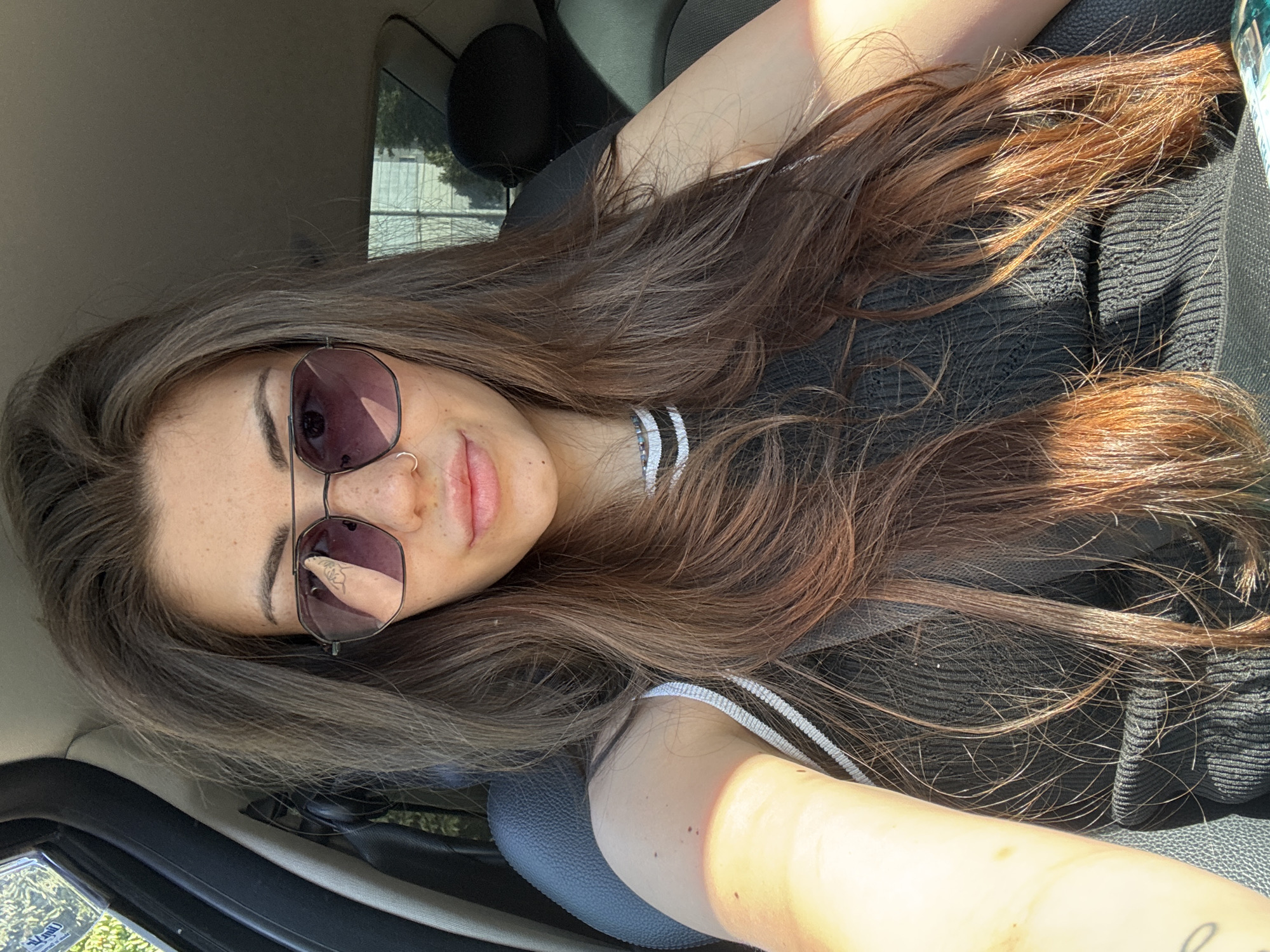 A young woman is pictured inside a car, likely parked, as evidenced by the stationary nature of the interior and the background view through the window. She is wearing sunglasses and a black top with white trim. The lighting suggests it is daytime, with sunlight casting shadows and highlights. Her hair is long and brown. The location is not identifiable as Sofia, Bulgaria from the visual information provided.