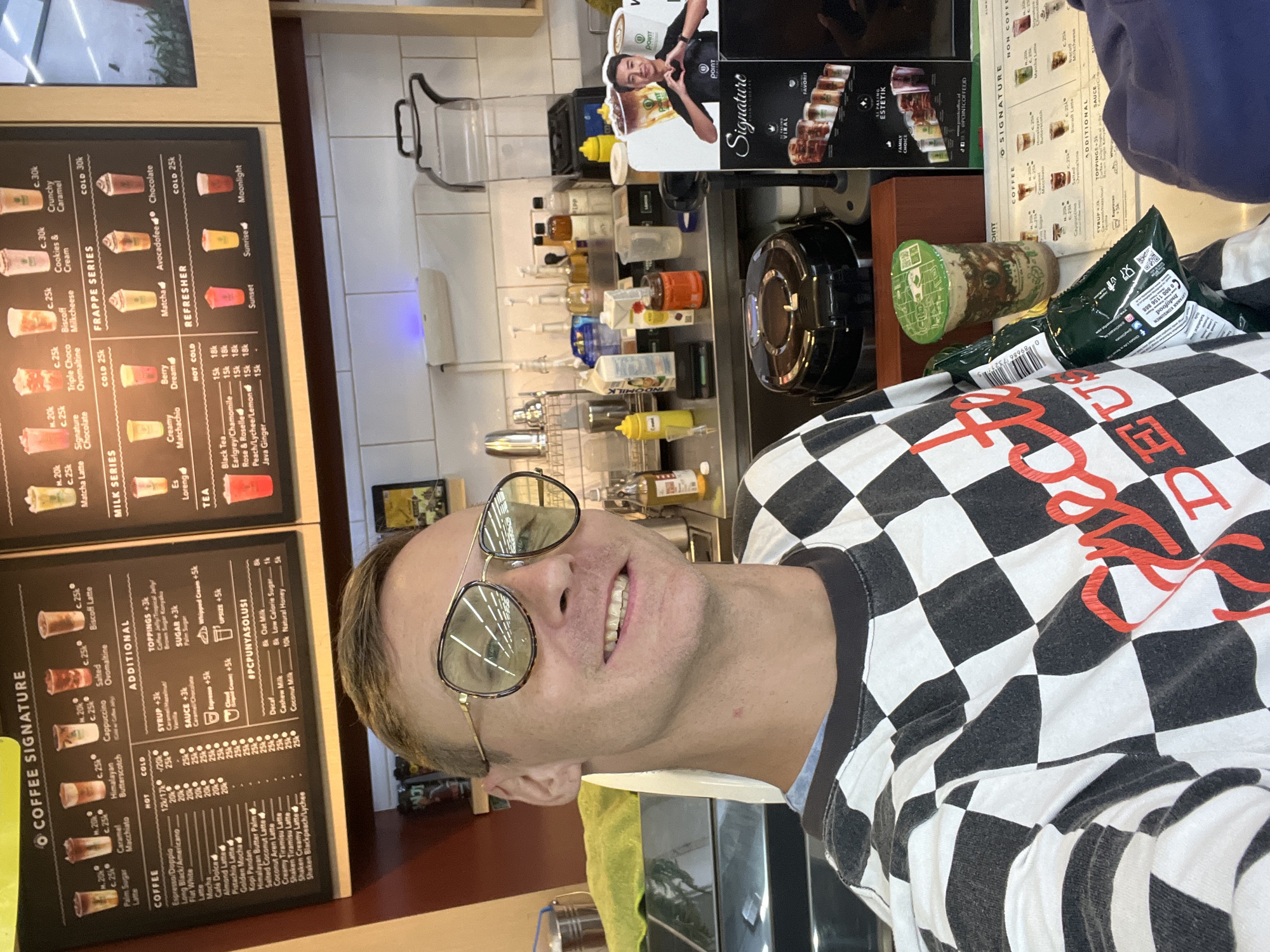 A young Caucasian male, wearing a black and white checkered long-sleeved shirt and aviator sunglasses, smiles at the camera in a selfie. He stands in front of a coffee shop counter, with a menu board displaying various coffee and frappe options in the background. The menu features item names such as "Palm Sugar Latte", "Himalayan Butter Palm", and "Cookies & Cream". To his right, a small cardboard cutout depicts a man making a heart shape with his hands, promoting "POINT COFFEE". Various coffee making equipment and ingredients are visible on the counter, including blenders, syrup bottles, and milk cartons. The environment appears to be a typical coffee shop setting in Sukawati, Indonesia.