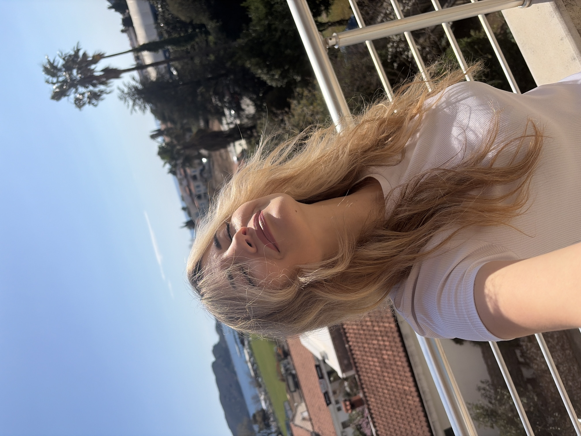 A woman with long blonde hair leans back against a white metal railing, with a view of Budva, Montenegro, in the background. The blue sky is visible above, and a palm tree is seen on the left side of the image. Behind the railing, red tile roofs of buildings can be seen. To the left of the roofs, a body of water is present, and beyond that, green areas and hills are visible. The woman wears a white top and has her eyes closed, facing upwards towards the sun.