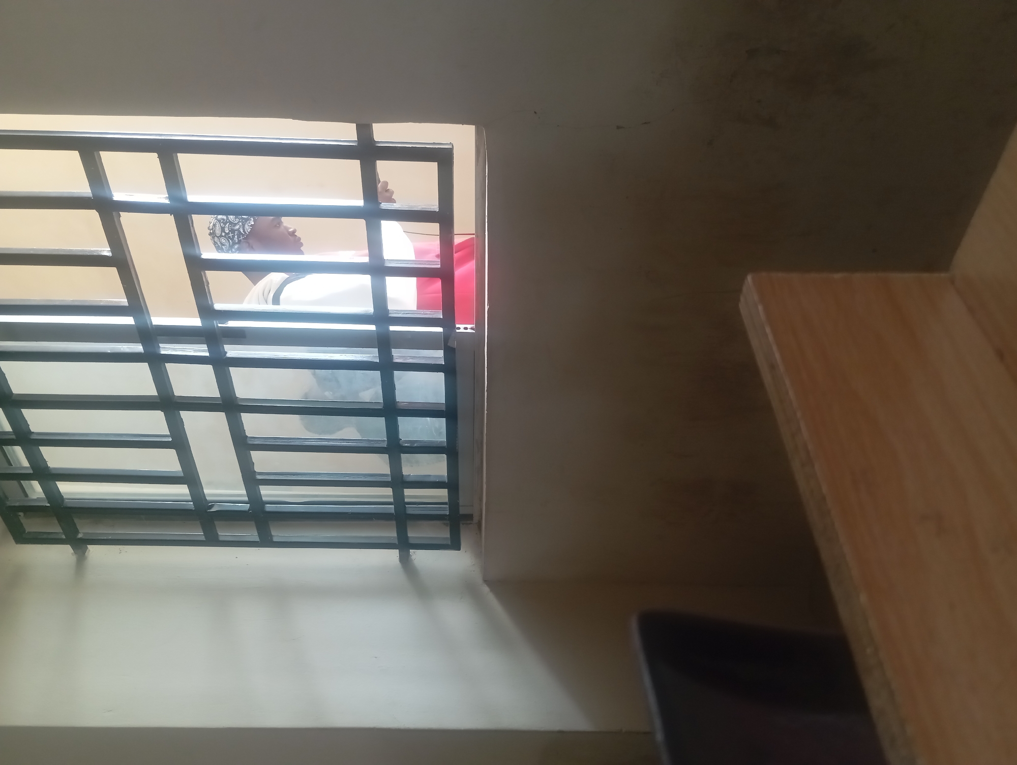 An interior shot captures a person in a room, viewed through a barred window. The person, whose face is partially obscured, wears a headscarf and appears to be looking down. They are dressed in white and red clothing. The room itself features neutral-colored walls and a barred window frame that extends across the upper portion of the image. In the foreground, a wooden surface and a dark chair are visible, suggesting a classroom or office setting. The lighting indicates natural light streaming in from the window.