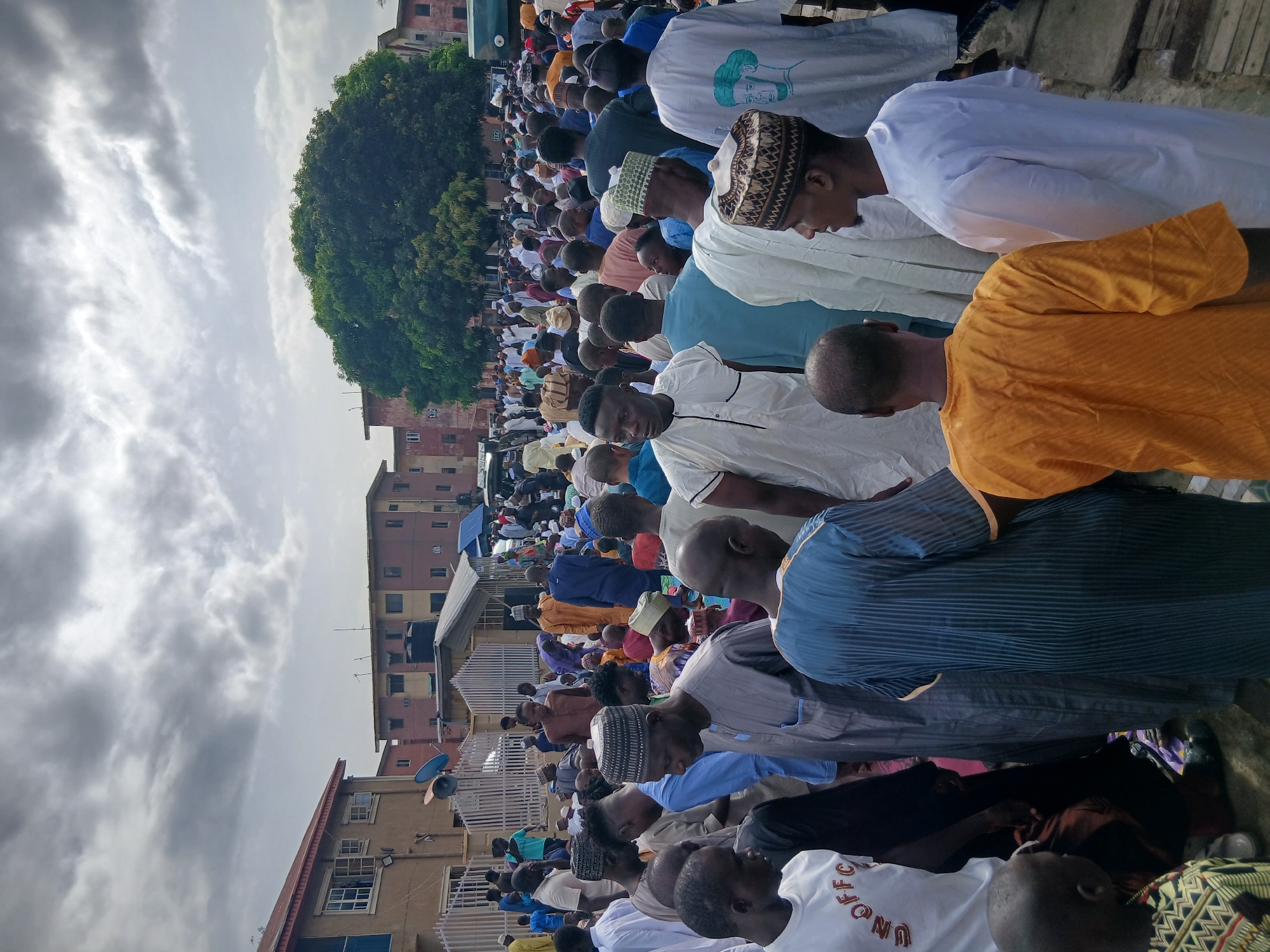 A large group of people are praying outdoors in Lagos, Nigeria. The majority of the people are men wearing traditional Muslim clothing. The prayer area is located in a somewhat urban setting, as indicated by buildings in the background, and a large tree. The sky is overcast.