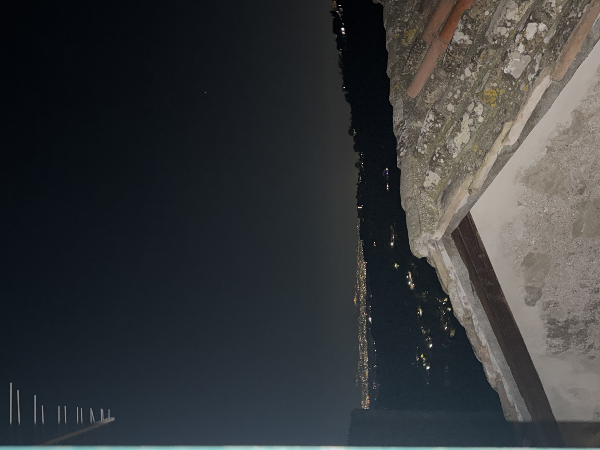 The image presents a night scene overlooking a body of water, potentially a lake or river. On the right, an aged stone structure, likely part of a building, is visible, featuring a decaying surface with exposed brick and mortar. In the background, on the opposite side of the water, a string of lights suggests a town or city stretching along the shoreline. The scene appears to be taken from a high vantage point, possibly from a building overlooking the water. The presence of the stone structure suggests an old or historic building in the vicinity. I cannot determine the specific location without additional information.