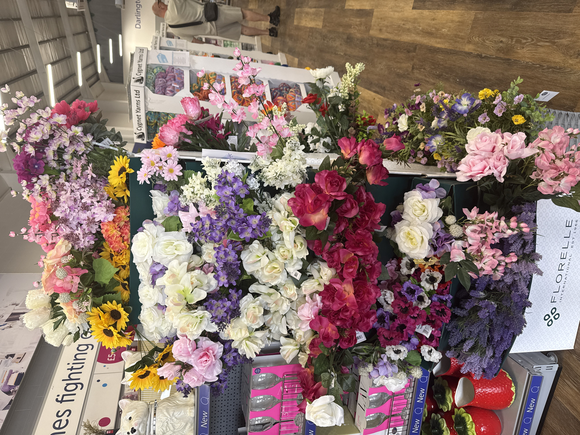 A medium shot captures a display of artificial flowers in a retail setting. The flowers are arranged in various clusters and hues, with prominent displays of pink roses, white roses, sunflowers, purple flowers, and lavender. The flowers are presented on shelves and in containers, with some items also visible behind the floral arrangements, including decorative ceramic strawberries and wine glasses. In the background, signage for "Cygnet Yarns Ltd" and "Darlington" is visible, indicating the location as Darlington, United Kingdom. A man wearing a light-colored t-shirt and shorts is seen in the background, browsing the shelves. The overall environment appears to be a well-lit store with wooden flooring.