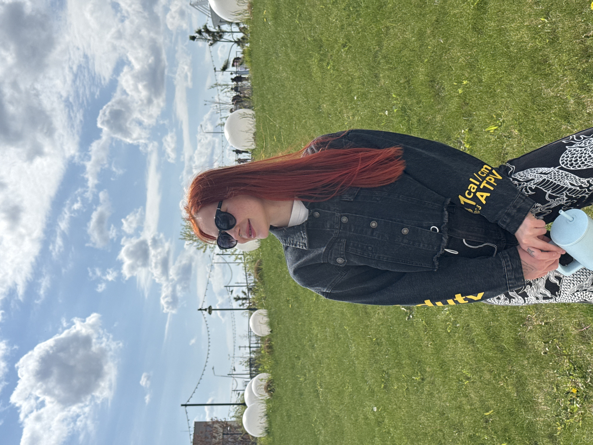 The image shows a young woman with red hair and sunglasses, seated on a grassy area with her hands clasped together. She's wearing a dark denim jacket with yellow lettering on the sleeves and black and white patterned pants. She is holding a blue water bottle. Behind her is a park-like setting with a bright blue sky and scattered white clouds. There are white sphere-shaped decorations, suggesting a public space. The location is Rostov-on-Don, Russia.