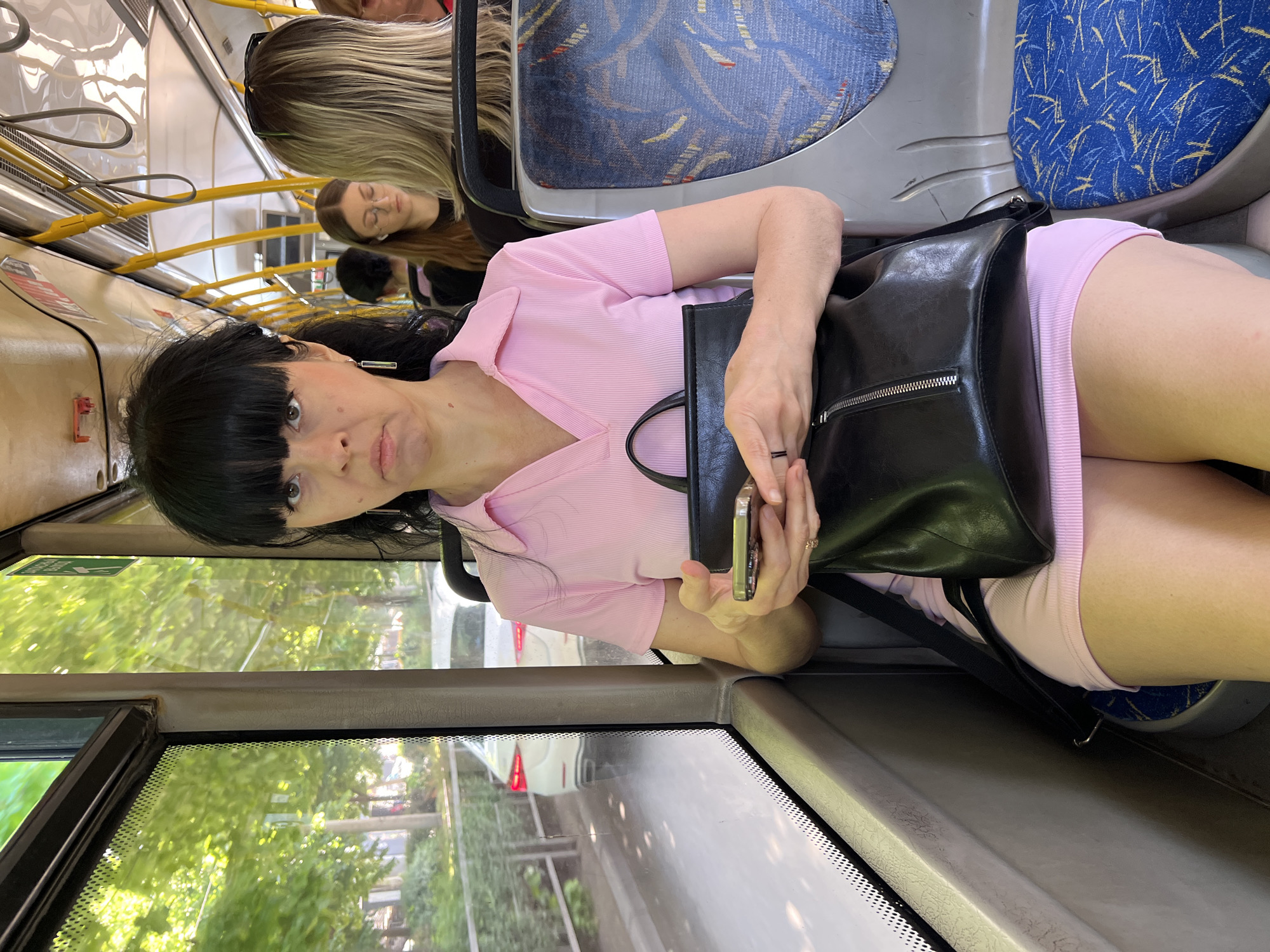 A medium shot depicts a woman with black hair and bangs on a bus, holding a smartphone and a black leather handbag. She is wearing a pink shirt and shorts. The bus interior is visible with yellow handrails and blue patterned seats. A window shows an exterior street with trees and buildings. The image location is Krasnodar, Russia. Other passengers are seated in the background. The woman is the primary subject of the image and is looking directly at the camera.