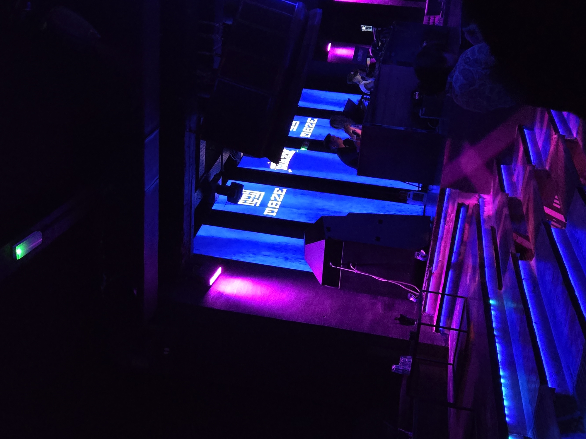 The interior is dimly lit with blue and purple neon lighting, creating a stark contrast. The walls are structured with a linear pattern of glowing neon strips. Visible text reads "MAZE." A group of people is present, though their activities are obscured by the darkness. A green light is discernible on the left side. The location is inferred to be Varna, Bulgaria, based on contextual data not evident in the image.