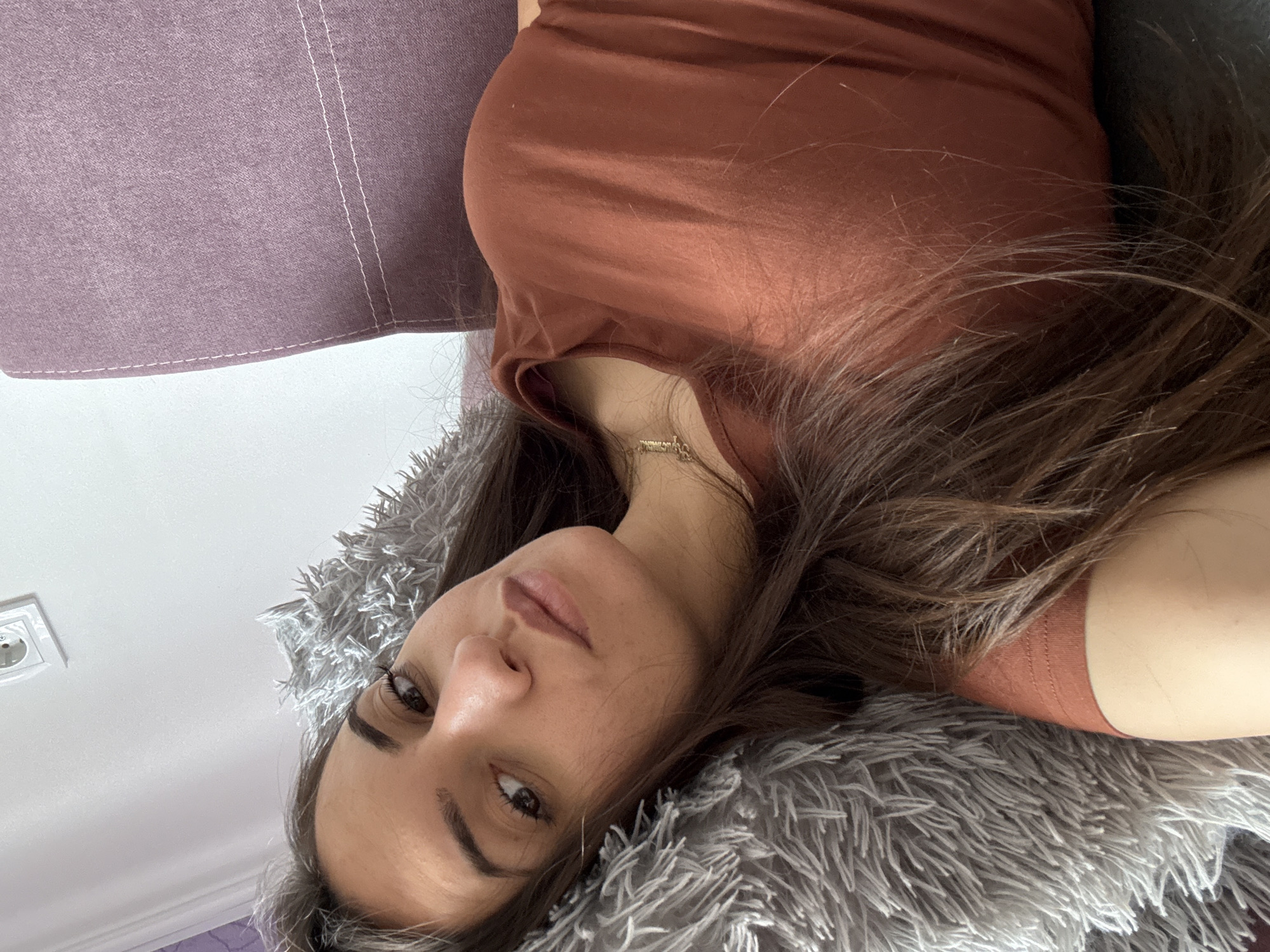 A woman with long brown hair is lying on a grey, furry pillow. She is wearing a brown t-shirt and a gold necklace. She is looking at the camera. The background includes a purple couch and a white wall with an electrical outlet. The location is not possible to determine from the information available in the image.