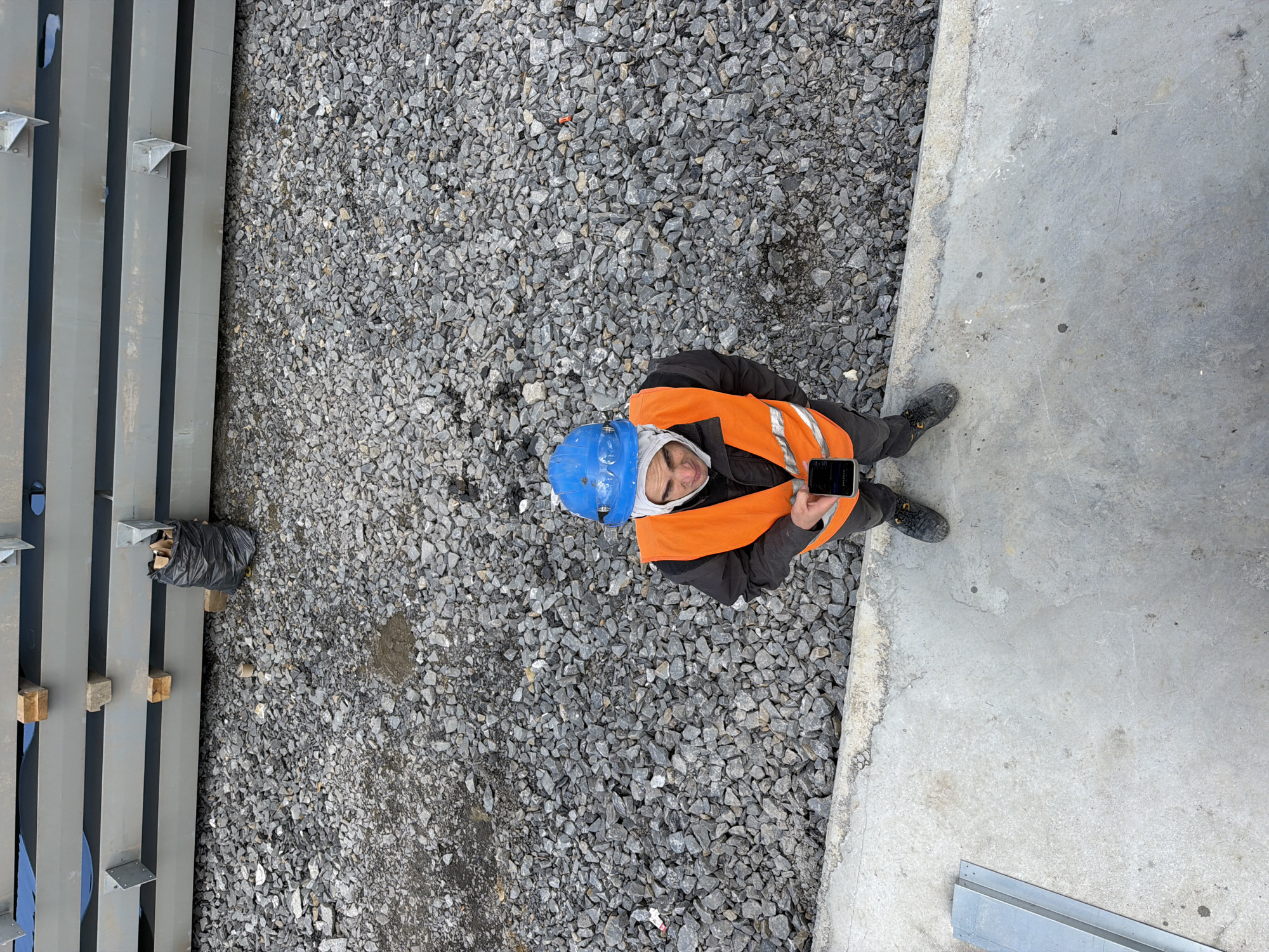 A construction worker stands on a concrete surface in Janowice, Poland. He is wearing a blue hard hat, an orange safety vest over a dark jacket, and dark pants. The worker is holding a smartphone in his hands, appearing to be looking at it. The environment is a construction site, with stacks of gray metal beams in the background, a pile of gravel covering the ground, and a black trash bag near the beams. A piece of metal construction material lies on the concrete in the foreground.