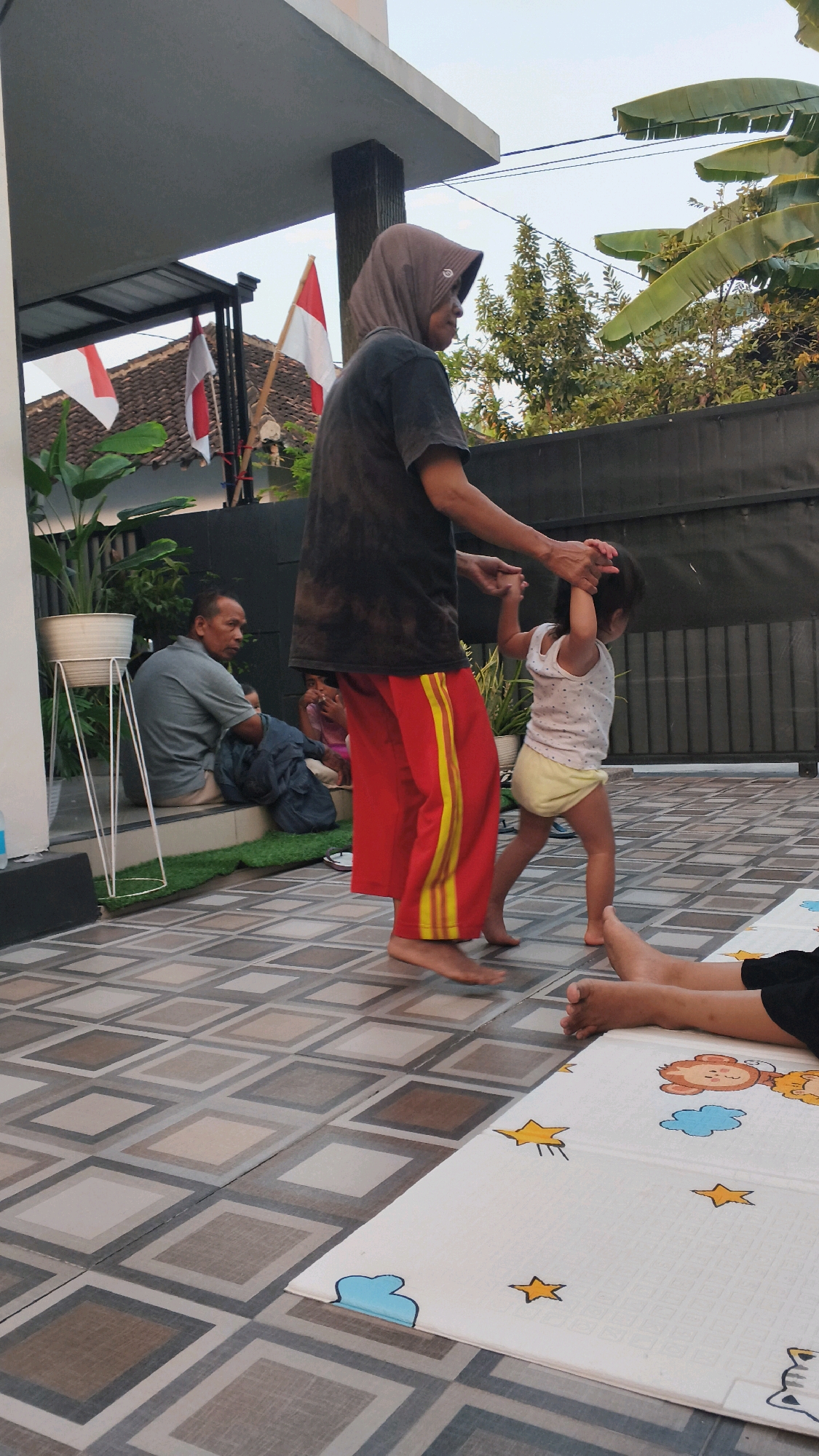 In Pundungsari, Indonesia, an outdoor scene unfolds on a tiled patio, possibly a residential backyard. A person wearing a headscarf and a dark t-shirt, along with red pants featuring yellow stripes, is holding the hands of a young child who is wearing a white sleeveless top and a yellow diaper. The child appears to be walking or dancing. In the background, a man in a grey shirt is sitting on a step next to a potted plant. Two Indonesian flags, red and white, are visible, along with some greenery and a dark fence. The sky is visible in the upper part of the image.