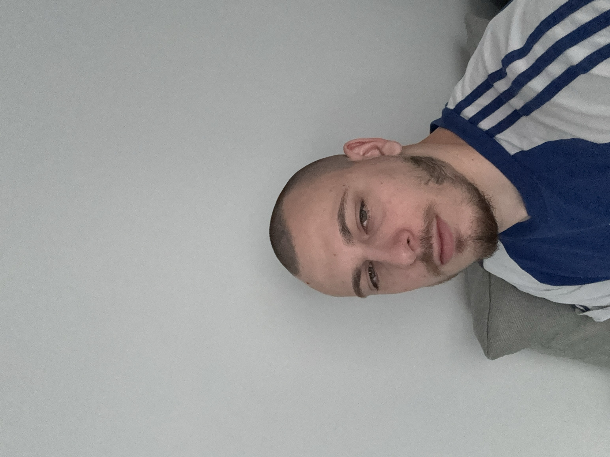 A man with a closely shaved head and a beard is visible in a rotated image. He is wearing a white shirt with blue accents and white stripes on the shoulders. A grey pillow is partially visible in the bottom right corner of the image. The background consists of a plain, light grey wall. The location is not identifiable from the provided image.