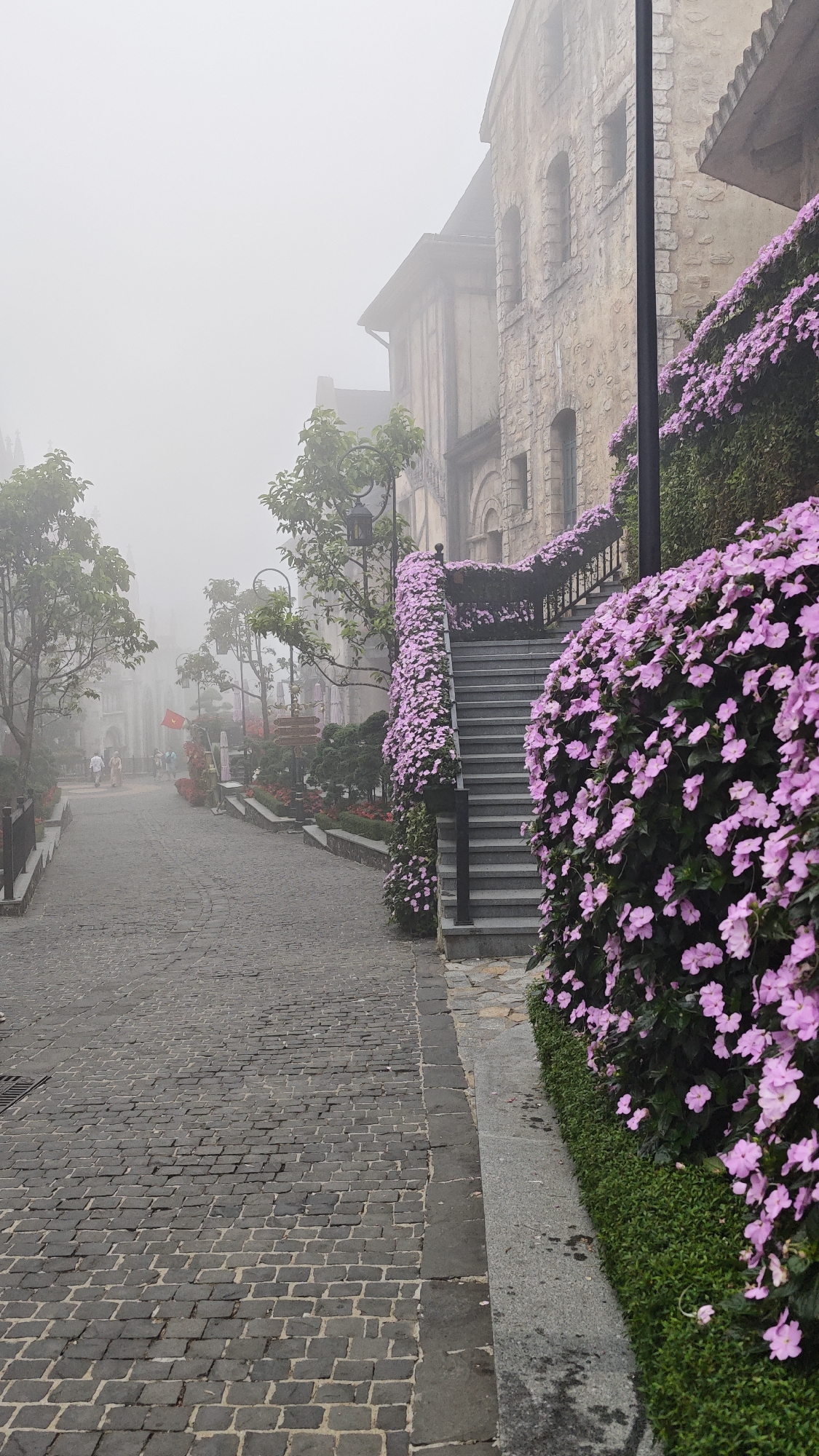 The image depicts a serene and atmospheric street scene, heavily enveloped in fog, giving it a dreamlike quality. The setting appears to be a European-style village or resort, likely in Ba Na Hills, Đà Nẵng, Vietnam, known for its themed architecture and frequent misty weather.
In the foreground and extending to the left, a dark gray cobbled street paves the way, curving gently into the distance where it is swallowed by the dense mist. On the right side of the frame, a prominent stone staircase ascends, its gray steps and dark railings almost entirely covered by a profusion of vibrant pink flowers that cascade down the sides. Lush green hedges line the base of the flowery display. Behind the staircase, a rustic building with light brown stone walls and arched windows is partially visible, its details softened by the fog. A dark, slender pole stands vertically near the building.
Along the left side of the cobbled path, various trees with green foliage emerge from the mist. Further down the street, decorative dark lampposts with intricate designs are spaced along the way. A wooden signpost with multiple arms can be seen, though no text is legible. Near the signpost, a small red flag, possibly a Vietnamese flag, is visible. A few indistinct figures of people are walking leisurely along the distant path, their forms blurred by the heavy fog.
The weather is extremely foggy, creating a soft, diffused light and a quiet, tranquil atmosphere. The time of day is not clearly discernible due to the overcast conditions, but the lack of harsh shadows suggests either early morning, late afternoon, or a heavily overcast day. The overall emotion conveyed is one of peacefulness, mystery, and charm, as the fog transforms the architectural and natural elements into a picturesque, ethereal landscape. No legible text is visible in the image.