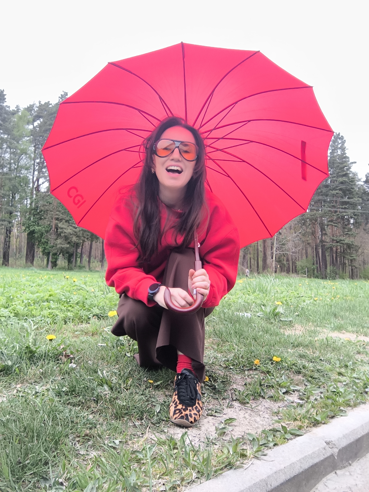 A young woman squats on a grassy area with dandelions, holding a red umbrella. The umbrella has a "CGI" label on it. She wears a red hoodie, brown pants, and leopard-print sneakers with red socks visible. She also has orange sunglasses and a smartwatch. Behind her are trees. A concrete curb is at the bottom right of the image. The location is a park in Riga, Latvia, based on the vegetation and architectural style common in the city.